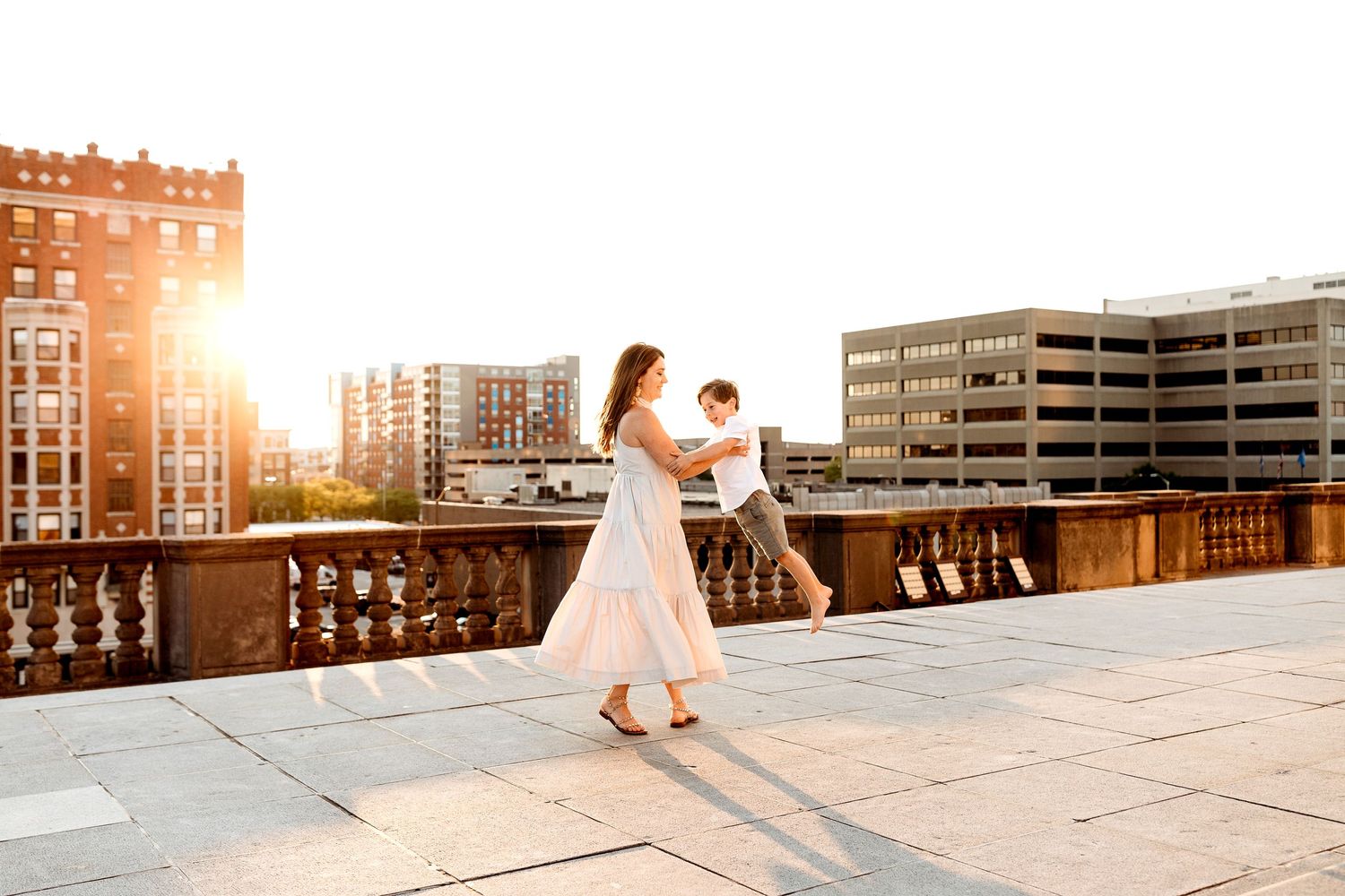 A woman in a white dress joyfully spins a child on a sunlit urban rooftop, with a warm sunset and tall buildings in the background
