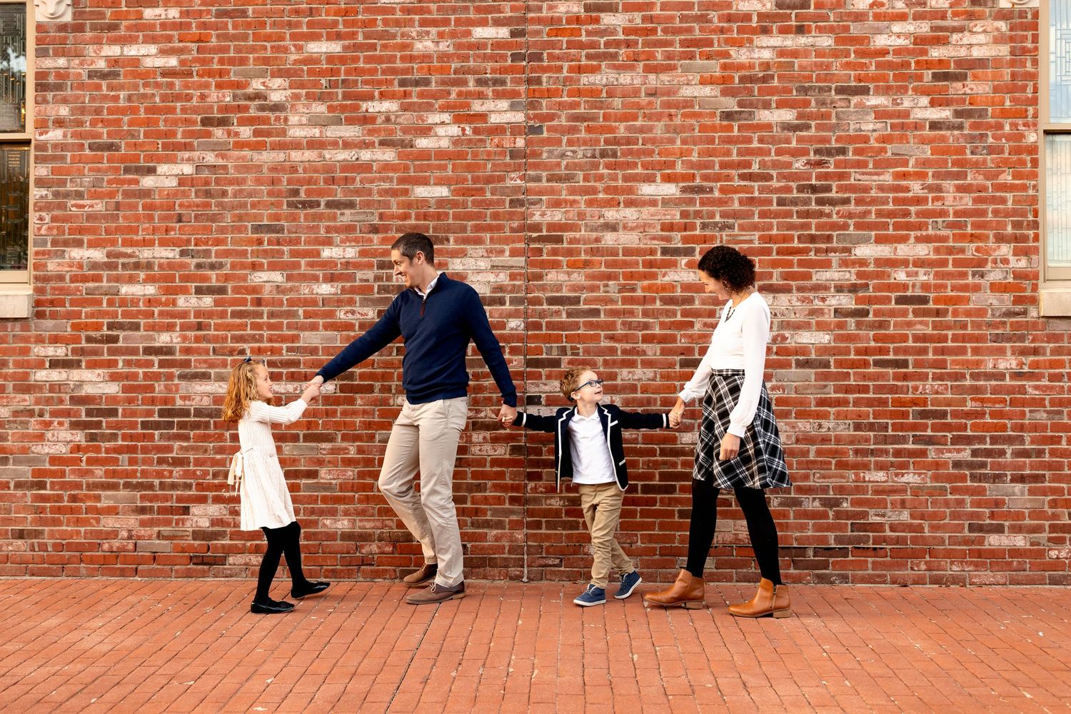 A family of four smiling, holding hands in front of a red brick wall for family portraits in Zionsville Indiana