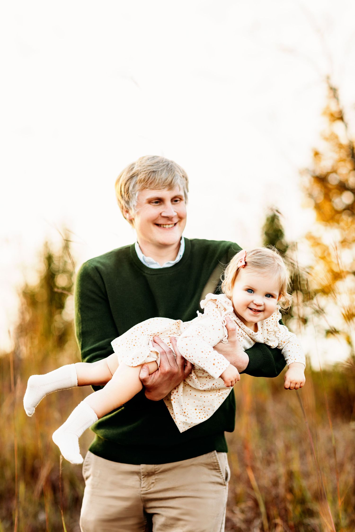 A person in a green sweater joyfully holds a smiling toddler wearing a floral dress in a sunny, autumnal field. Both appear happy