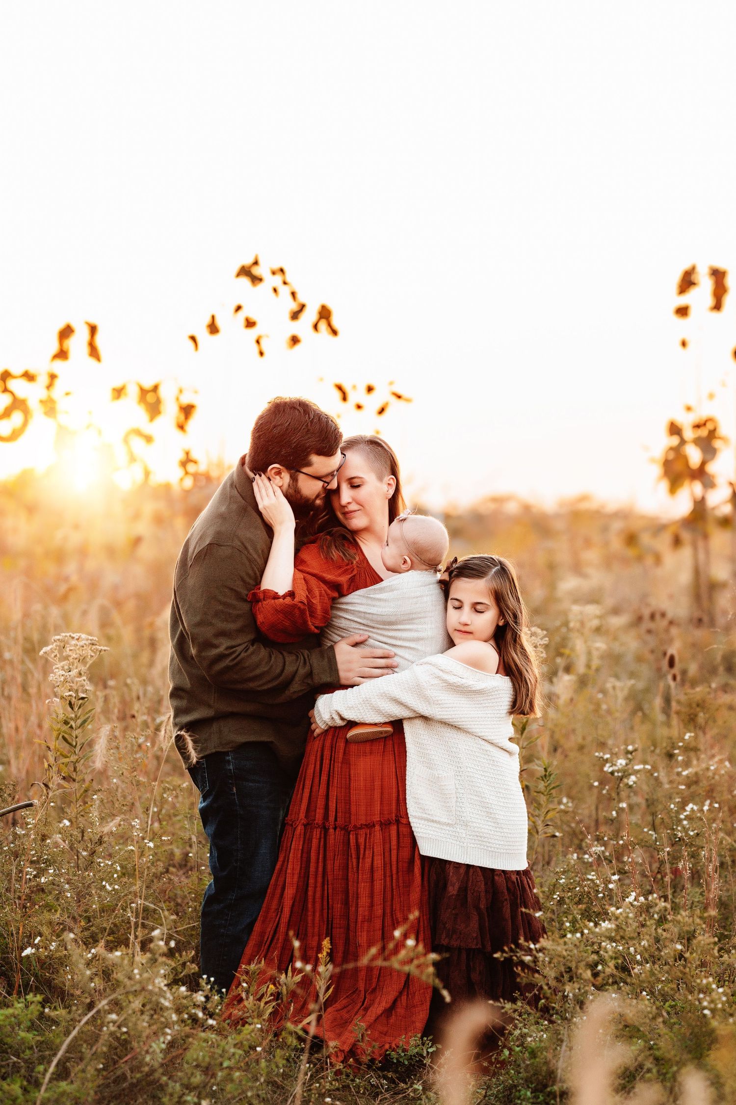 A family stands in a sunlit field at sunset. Parents embrace, holding a baby, while a young girl leans against them, conveying warmth