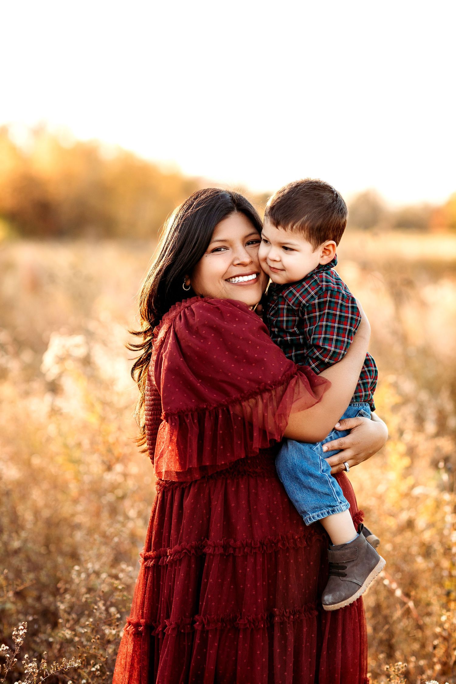 A smiling woman in a red dress holds a young boy in a plaid shirt in a sunlit field, conveying warmth and affection.
