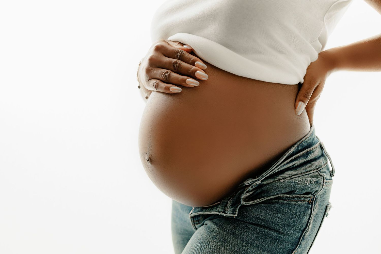 Pregnant woman in a white crop top and jeans, gently holding her bare belly with one hand, symbolizing maternal care.