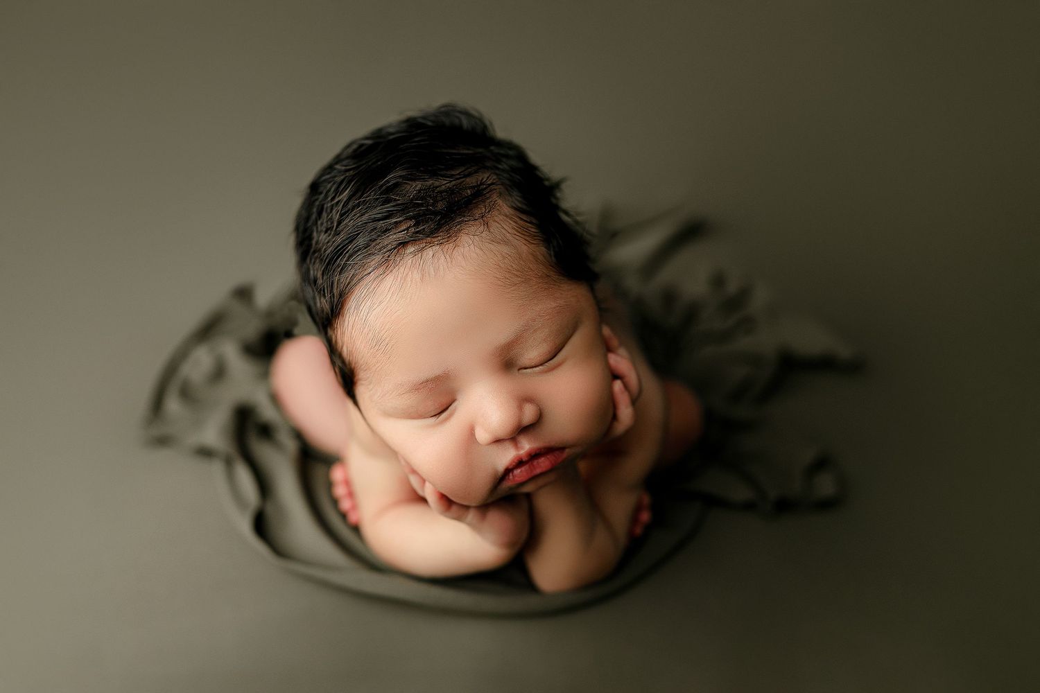 Sleeping newborn with dark hair rests with cheeks on hands, set against a soft, dark green background. The scene conveys peace and innocence