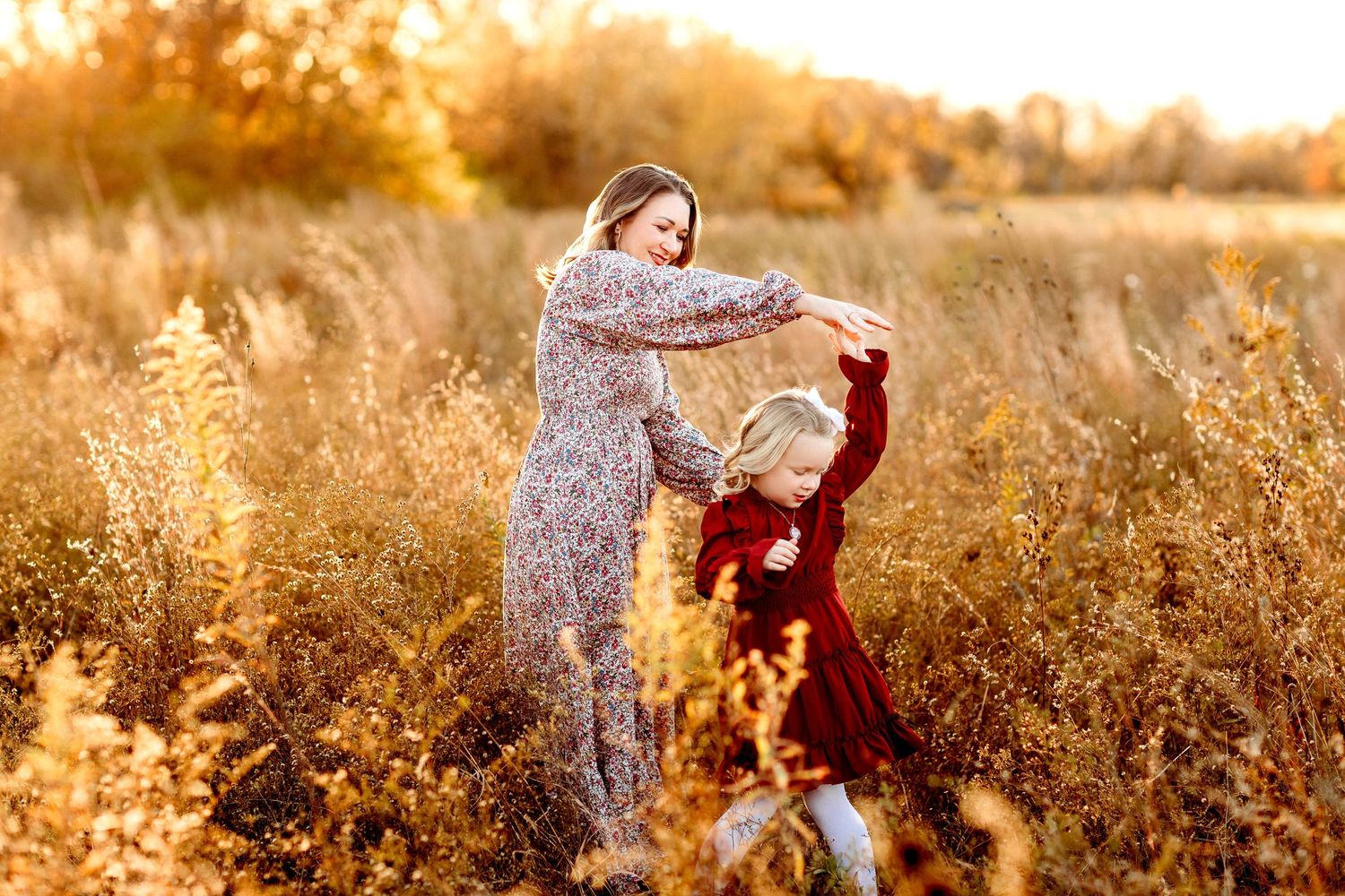 A mother and daughter dance in a sunlit autumn field, surrounded by tall golden grasses. The warm tones evoke a feeling of joy