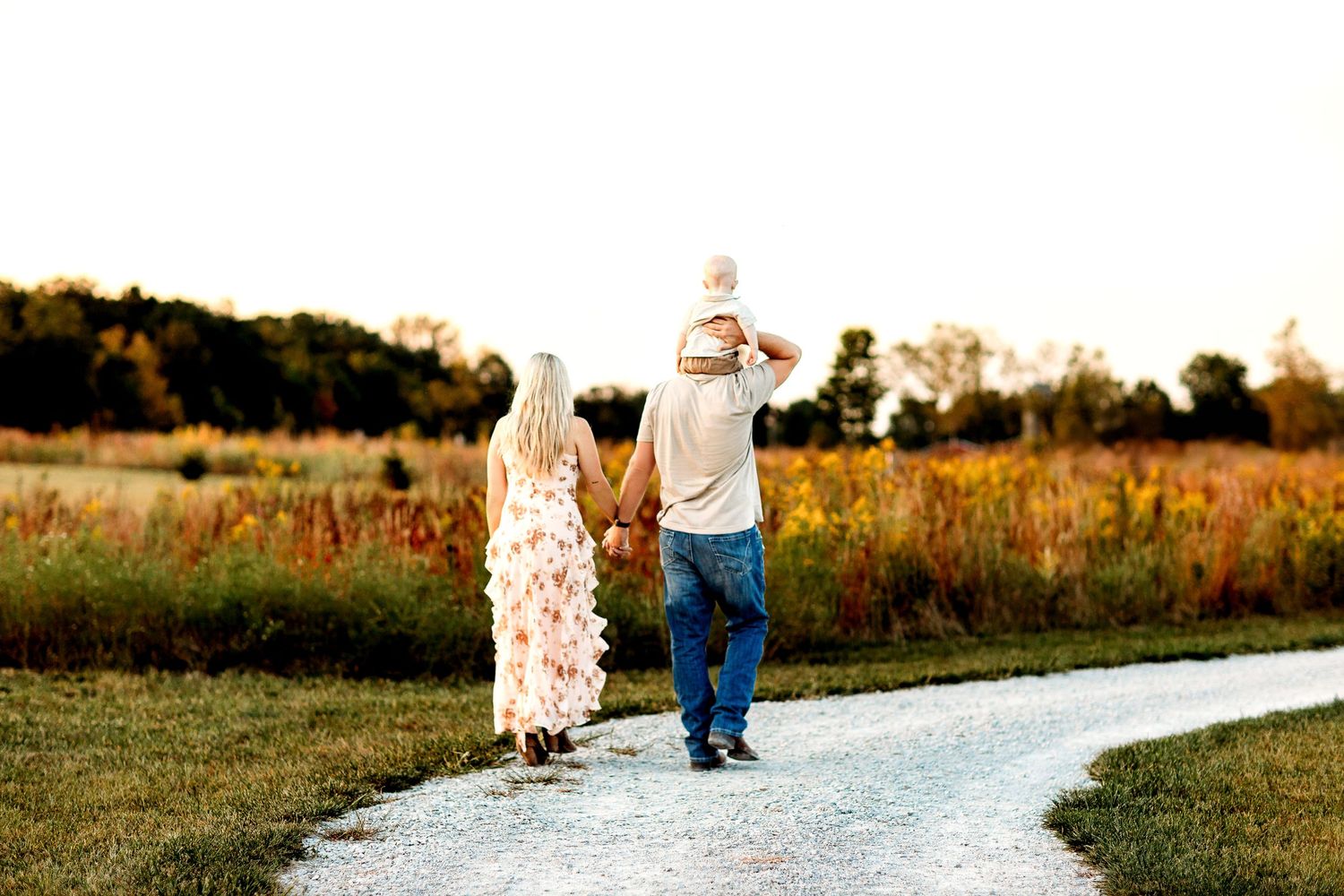 A family of three walks down a winding path through a scenic field. The woman in a floral dress, the man carries a child on his shoulders.