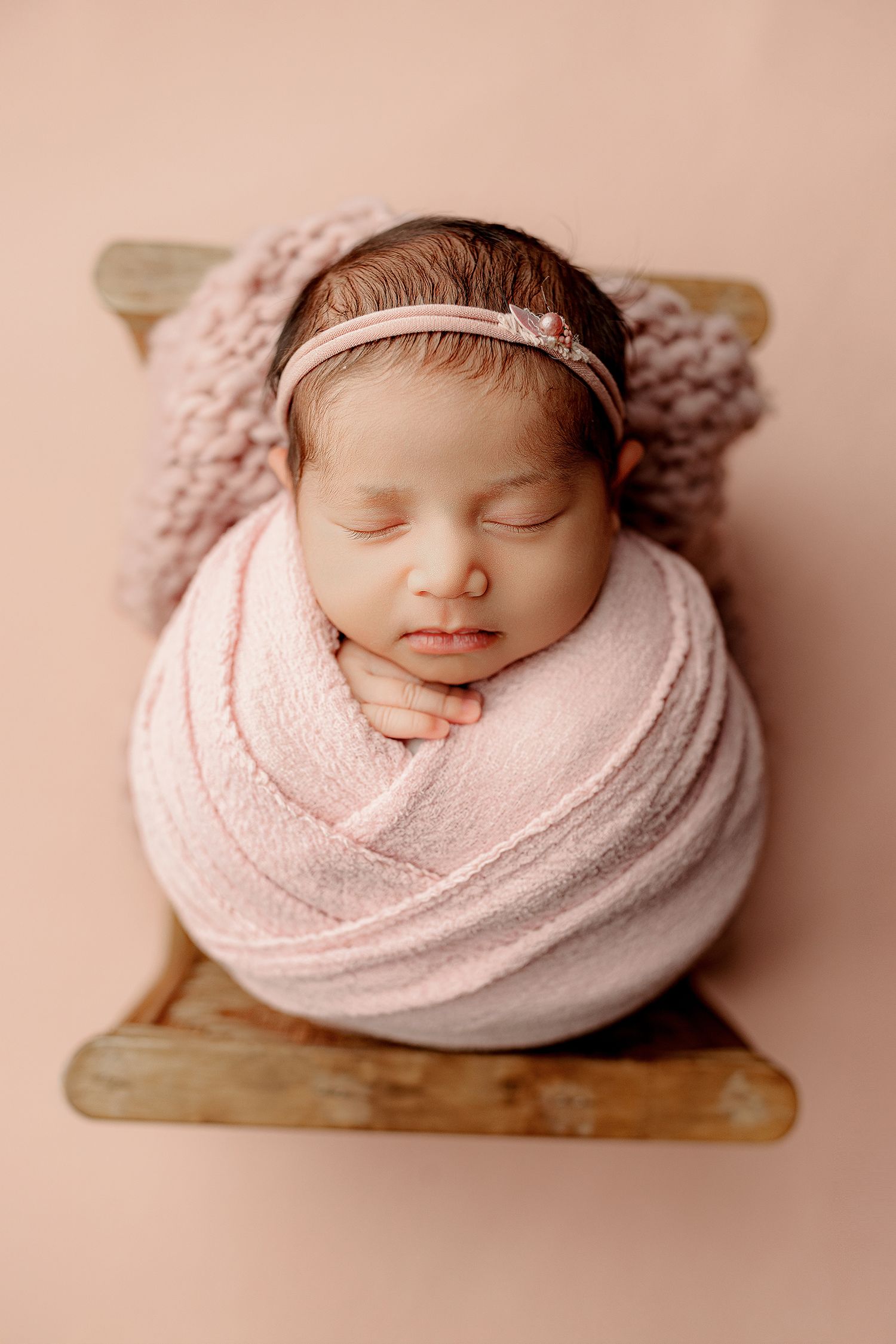 A sleeping baby, swaddled in soft pink fabric, rests peacefully on a small wooden bed.
