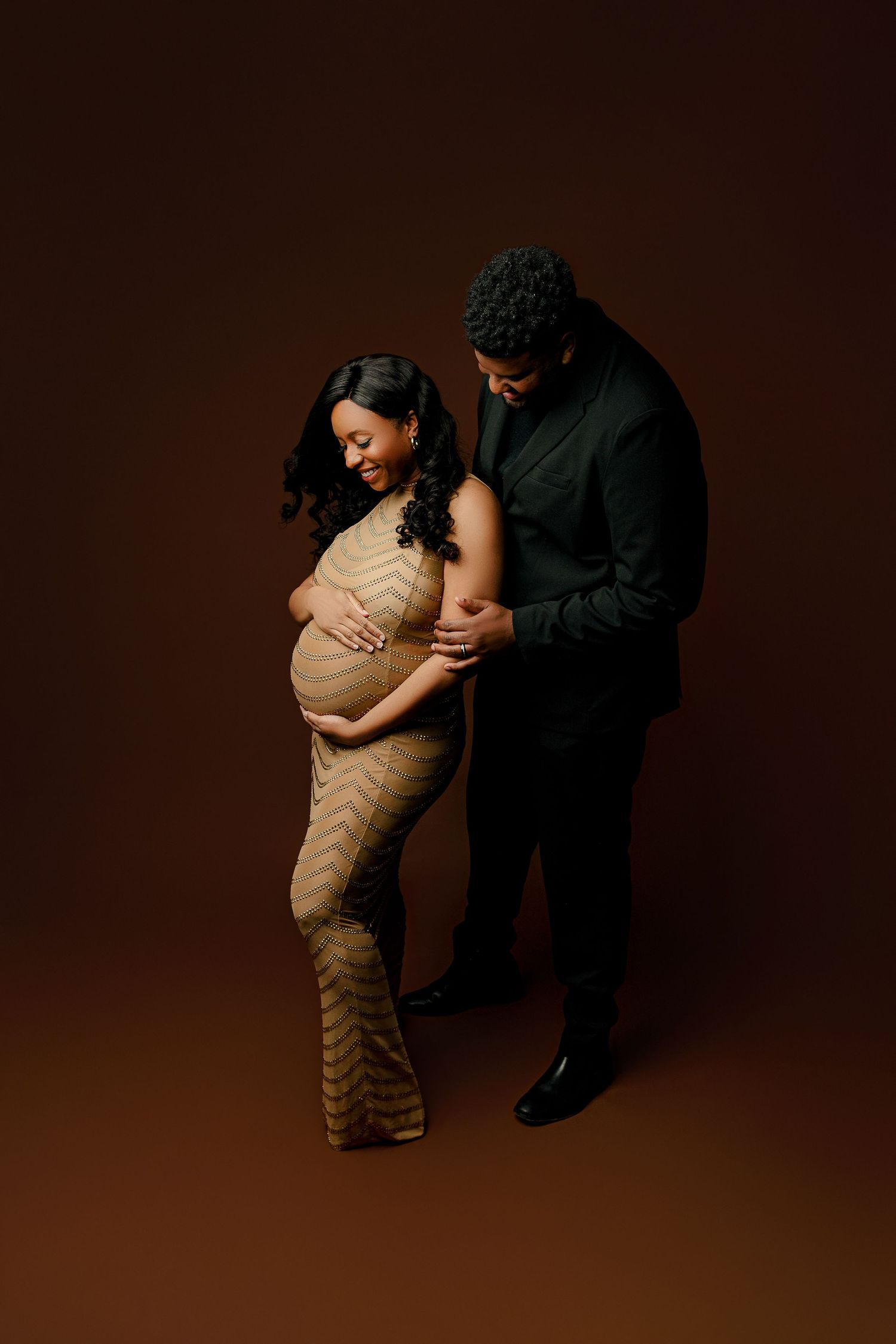 A pregnant woman in a gold dress stands smiling, cradling her belly. Beside her, a man in black gently embraces her against a brown drop