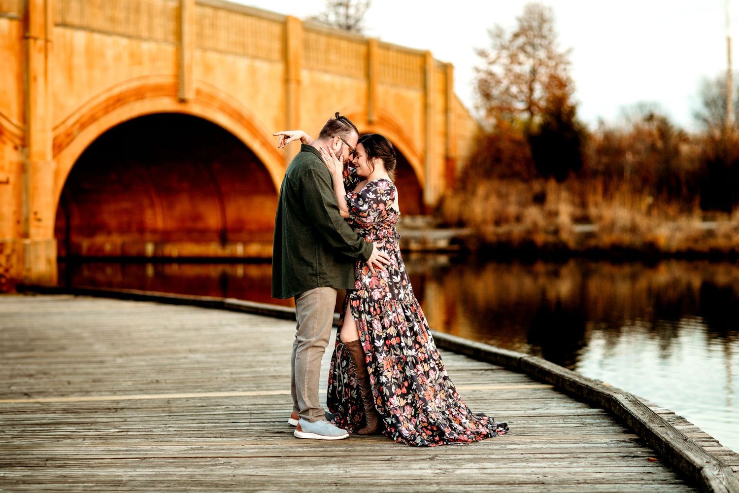 A couple embraces on a wooden walkway by a calm water. They are near an arched bridge. The mood is romantic and serene