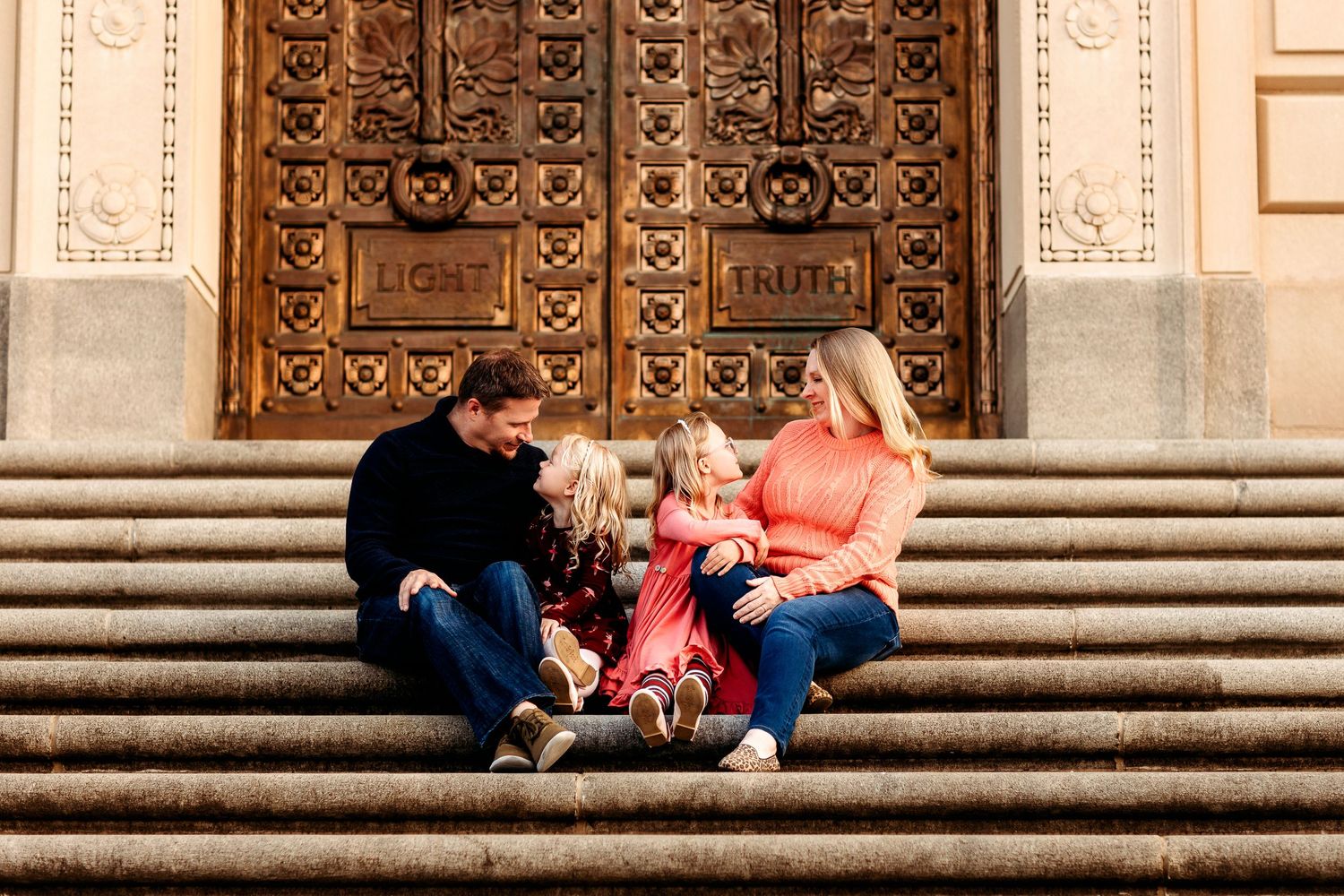 A family of four sits on steps before an ornate door at Indiana War Memorial. Parents smile at their young daughters, all in casual clothing