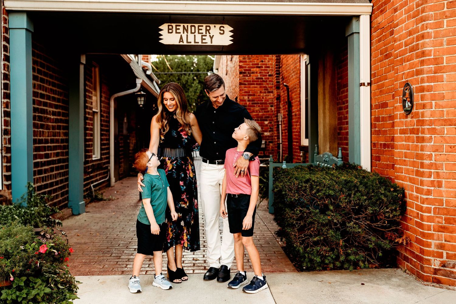 A family of four stands smiling in Bender's Alley in Zionsville Indiana The parents wear casual clothes, and the children look up smiling
