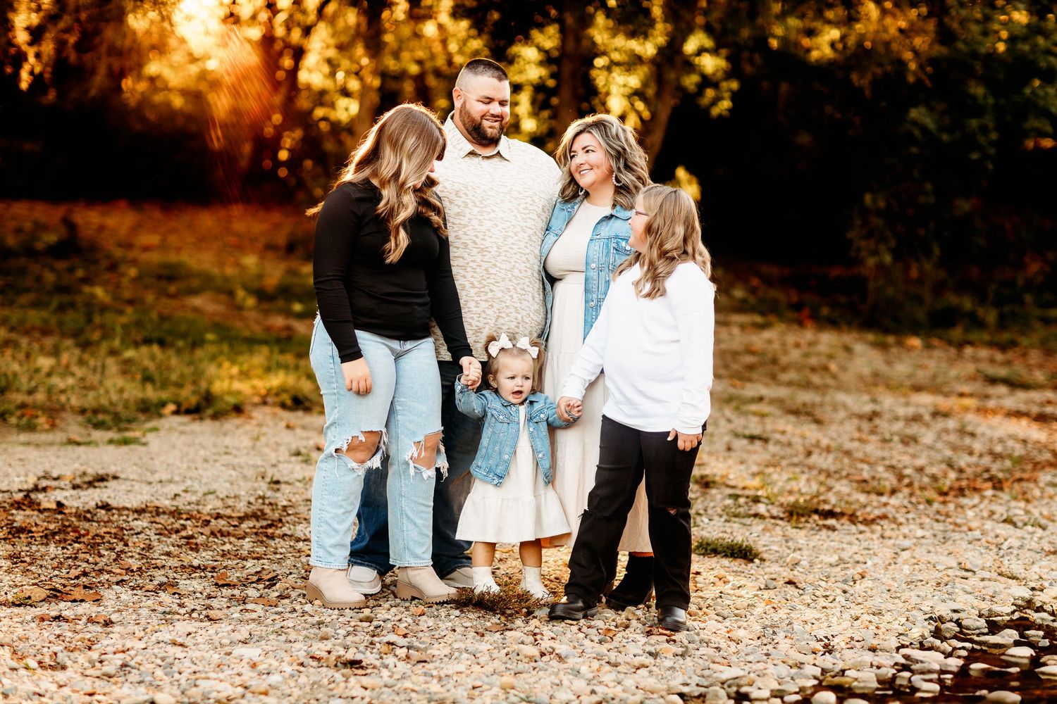 A family of five stands on a pebbled path amid autumn trees. They smile warmly, conveying a sense of togetherness
