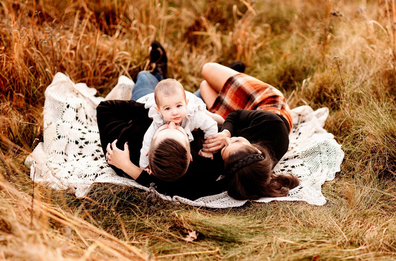 A family of three lies on a blanket in a field, with the baby in the center. The warm, autumn colors create a cozy scene