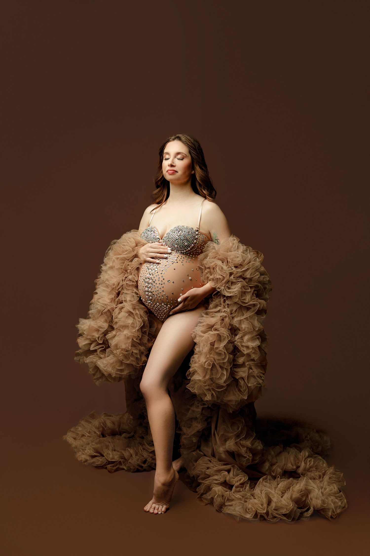 Pregnant woman in a jeweled bra cradles her belly, wearing a flowing brown tulle robe.