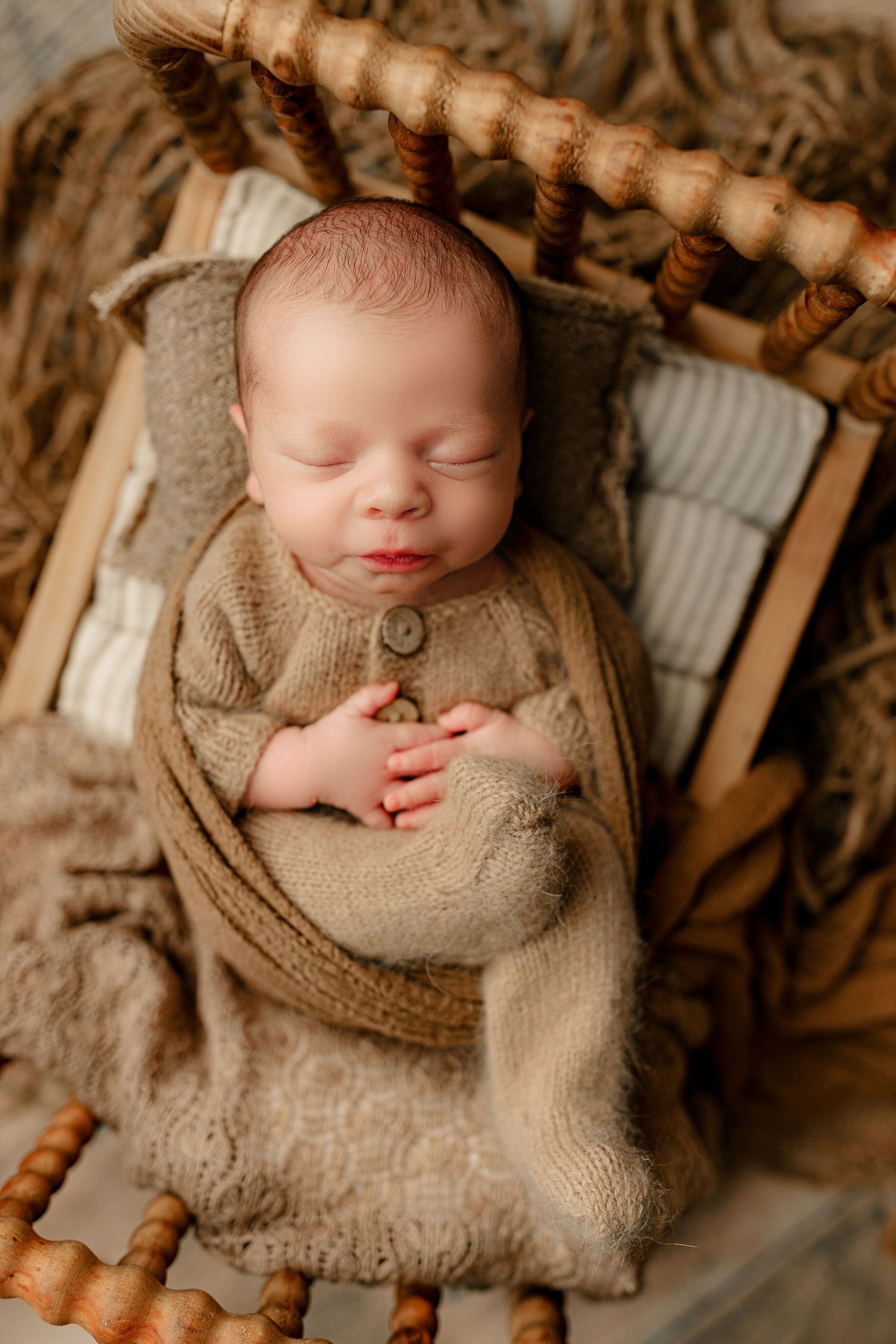 A peacefully sleeping baby in a cozy brown knitted outfit is swaddled on a rustic wooden chair. The scene conveys warmth and tranquility.