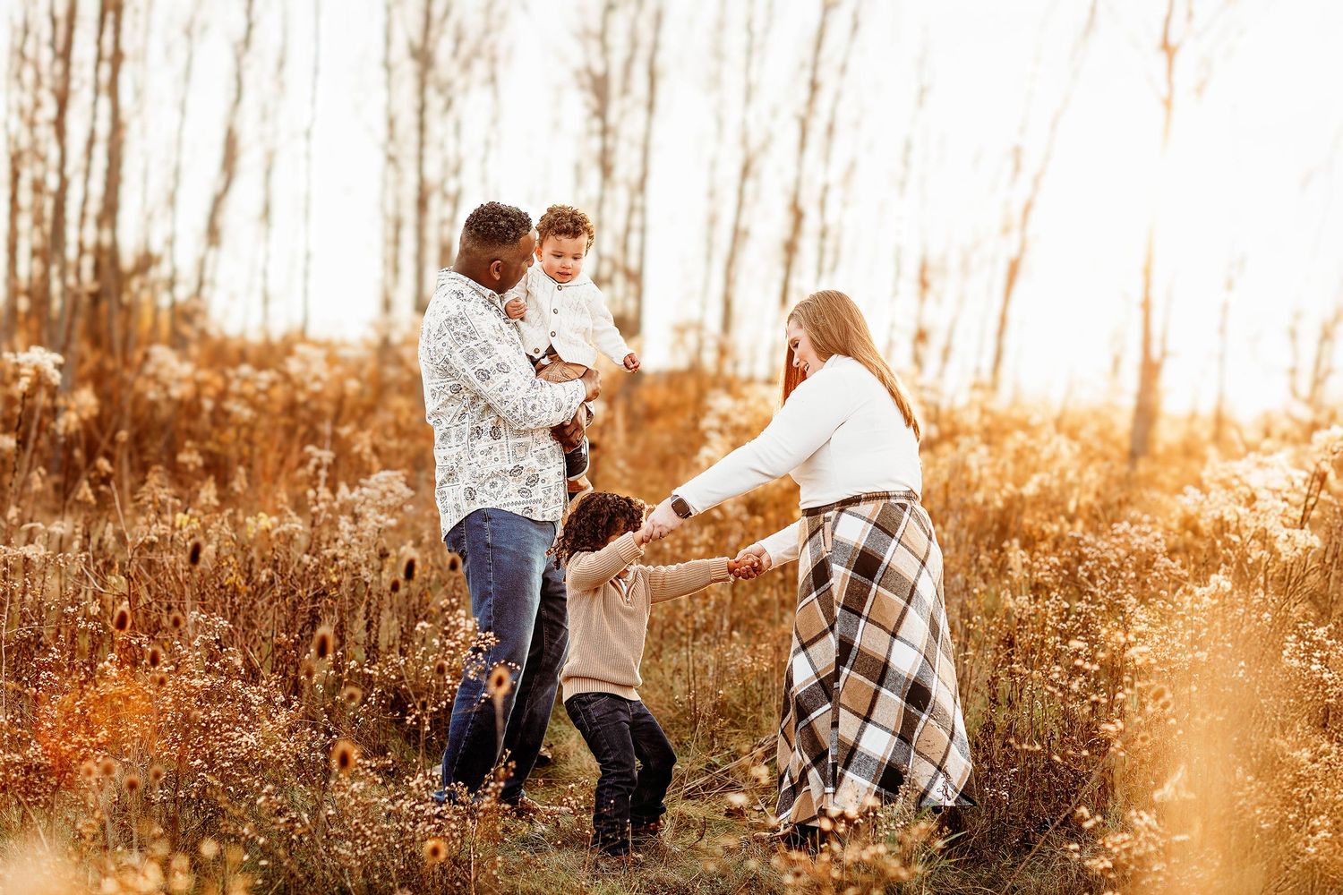 A family of four enjoys a sunny day in a field. Parents play with their two children, surrounded by tall grass and wildflowers