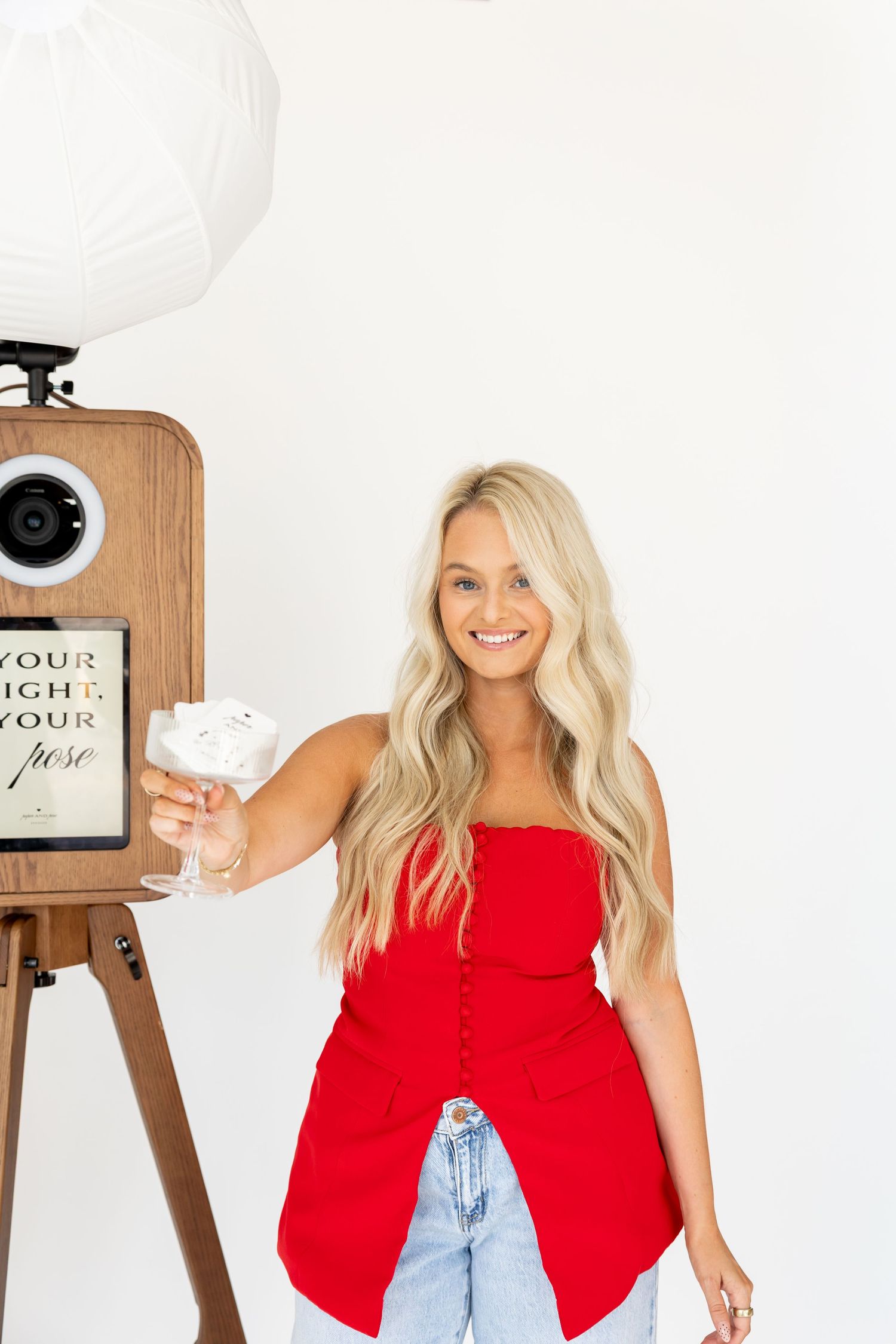 Woman in red top and jeans posing with vintage camera setup against white background.