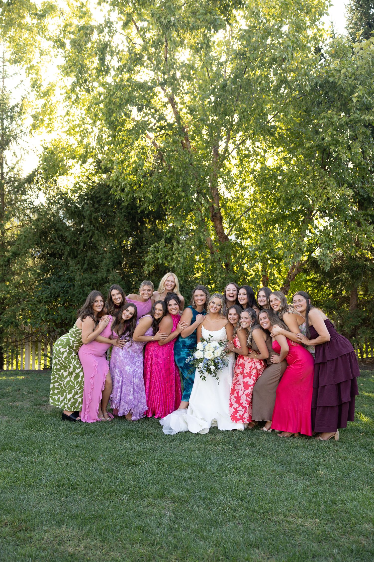 Wedding party in colorful dresses posed together on grass under trees.