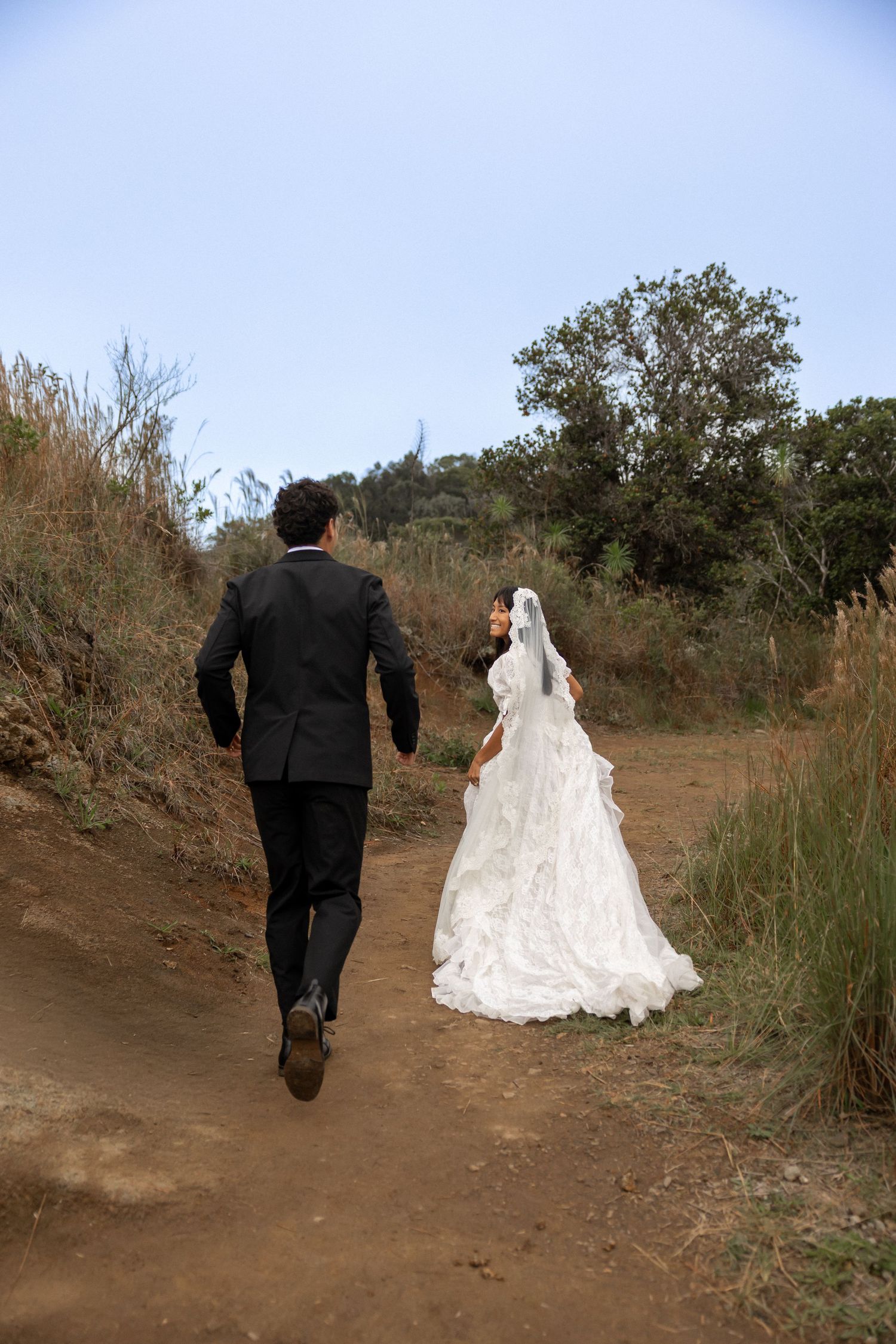 A bride and groom walking down a dirt path surrounded by nature after their wedding ceremony.