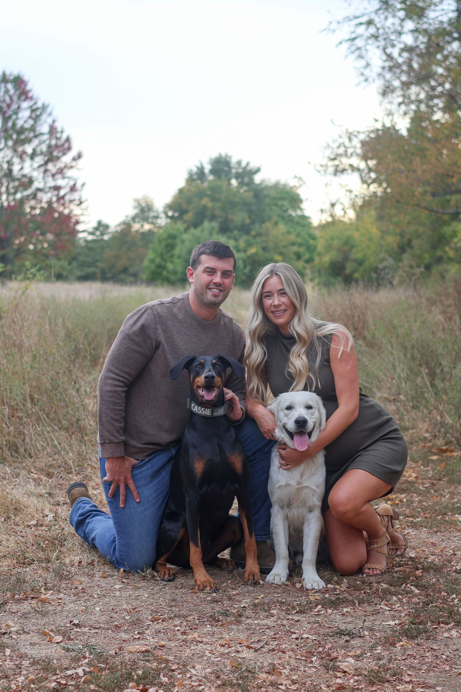 Happy couple sitting with their two dogs on autumn ground during outdoor photo session.