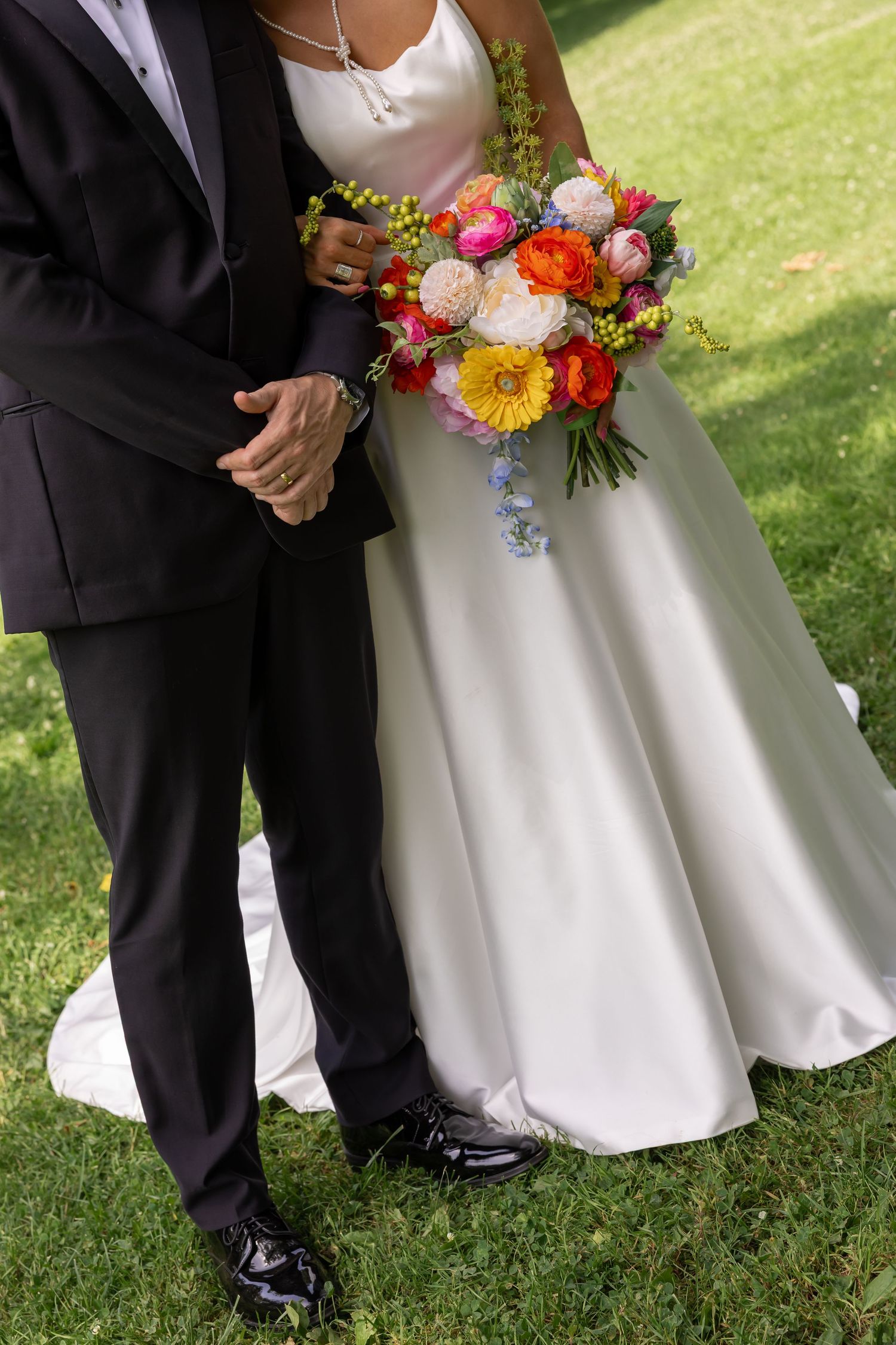 Bride in white gown holds colorful bouquet of flowers while standing with groom in black tuxedo on green grass.