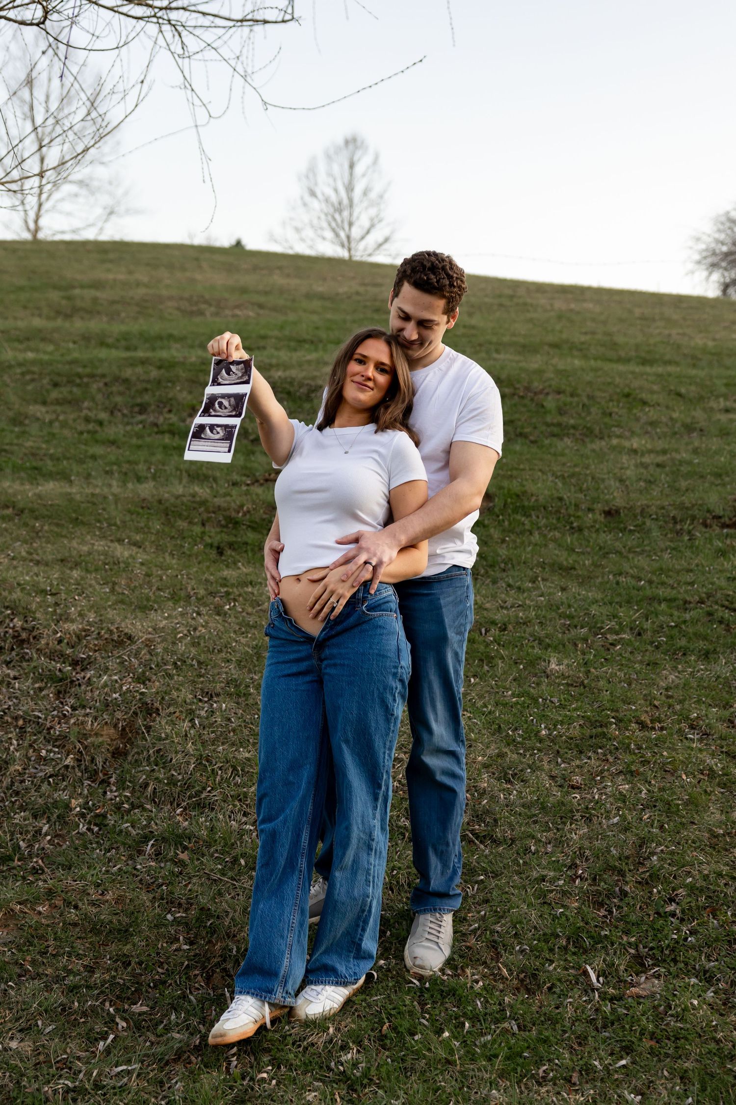 Happy couple in white tees and jeans holding ultrasound photos outdoors on a grassy hill, pregnancy announcement.
