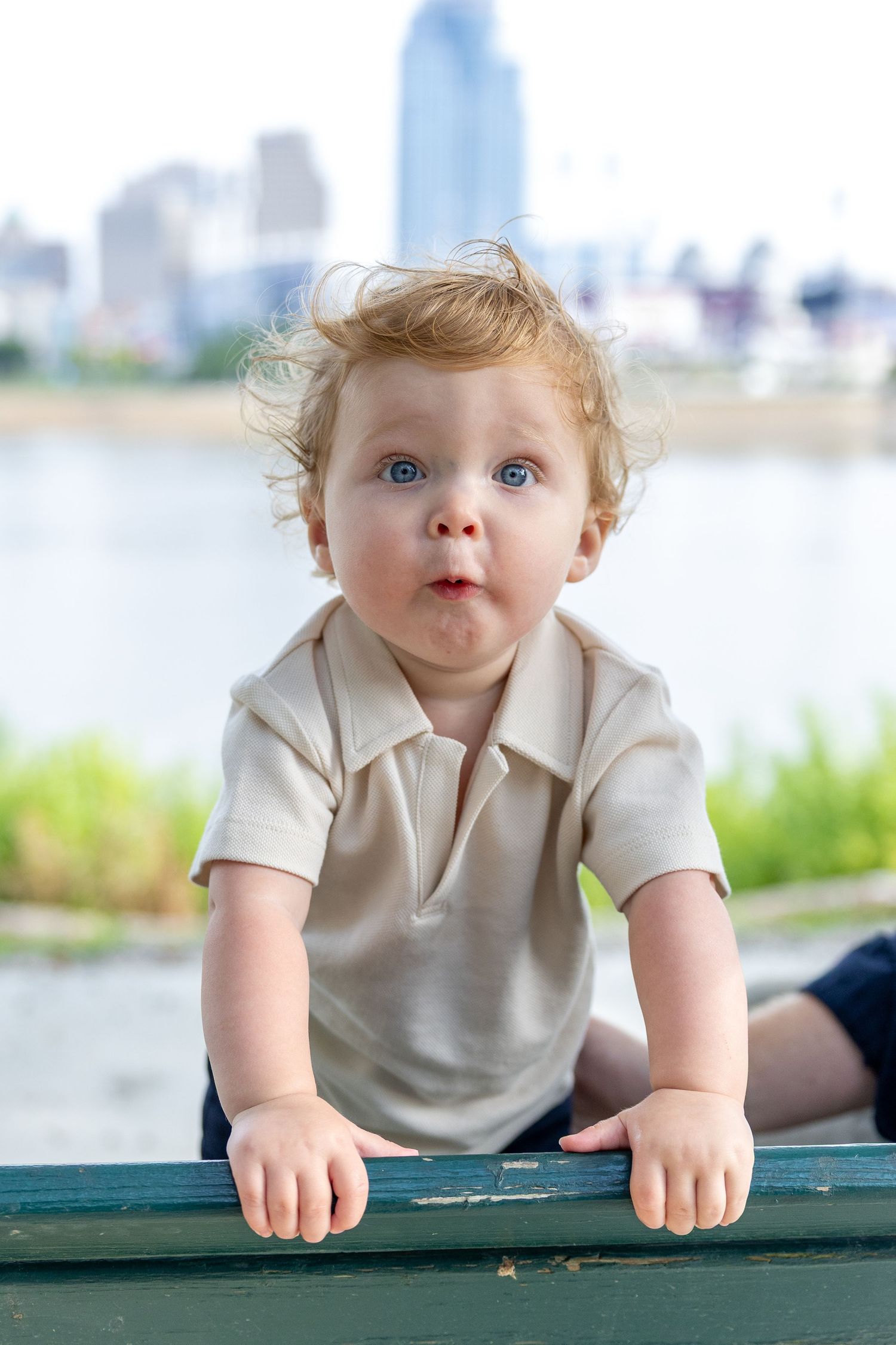 Baby in light colored outfit peering over edge with city skyline in background.