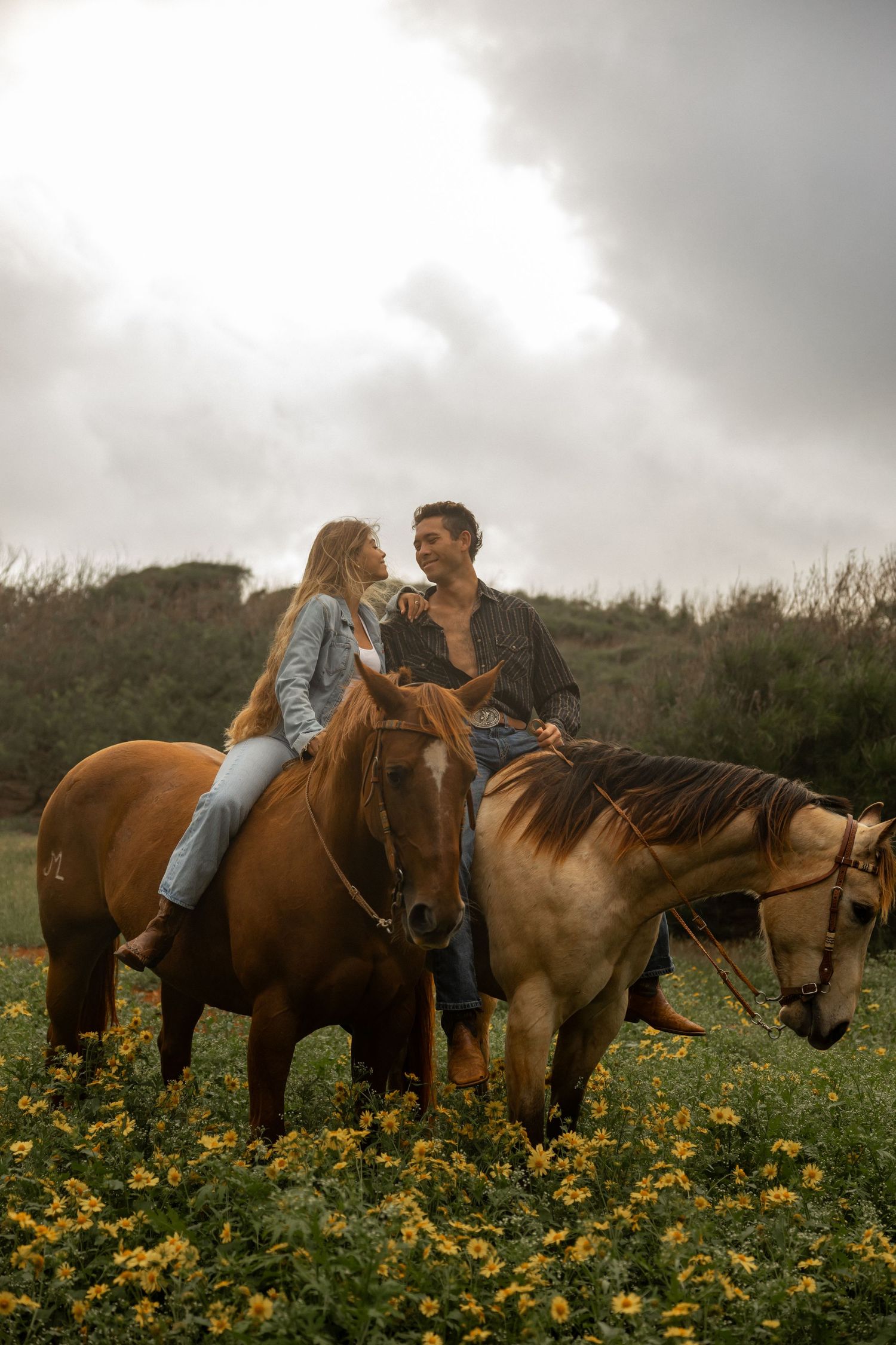 Couple sitting together on horses in a field of yellow wildflowers at sunset.