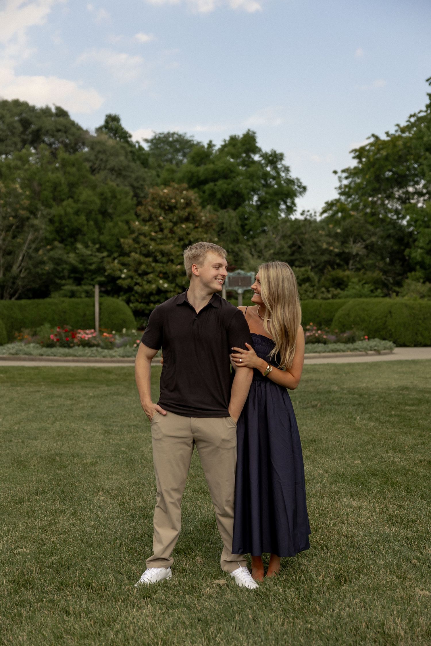 A couple smiles at each other while embracing on a green lawn, man in black polo and khakis, woman in navy dress.