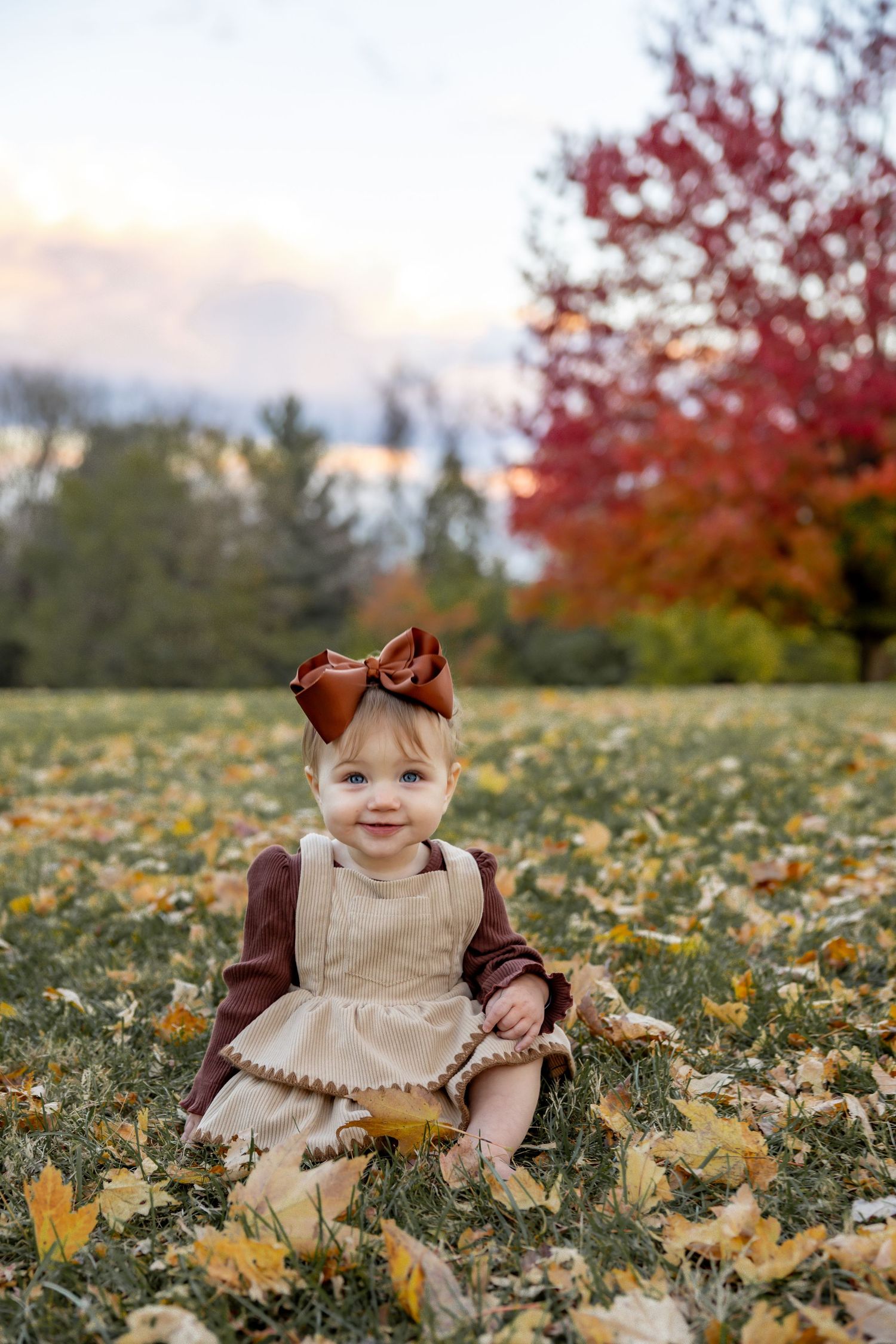 Baby sitting in fallen autumn leaves wearing a beige dress with brown bow.