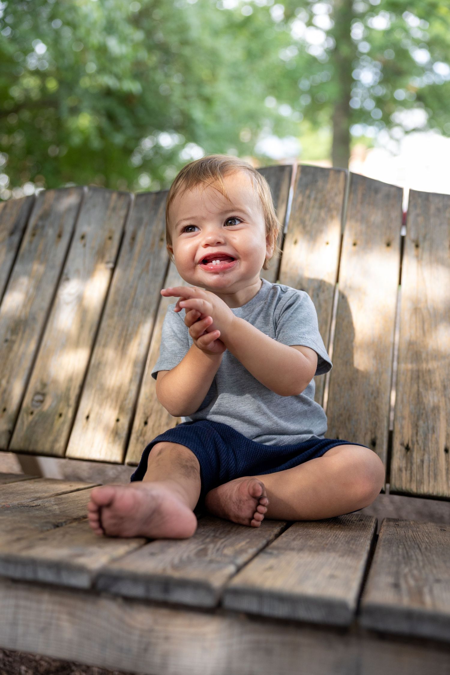 Young child sits playfully on wooden deck boards in outdoor setting.