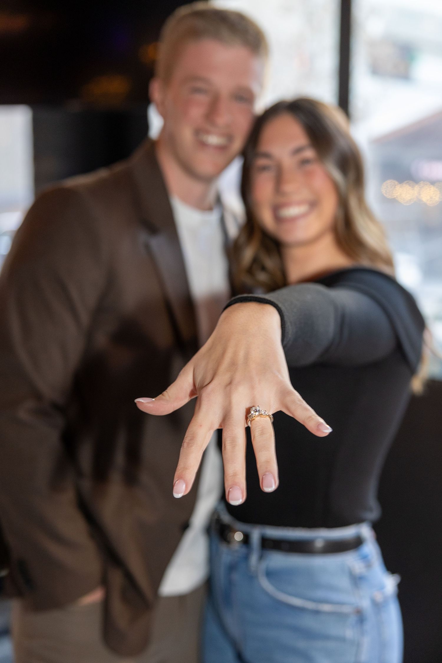 Close-up of engagement ring being shown with couple embracing in background.