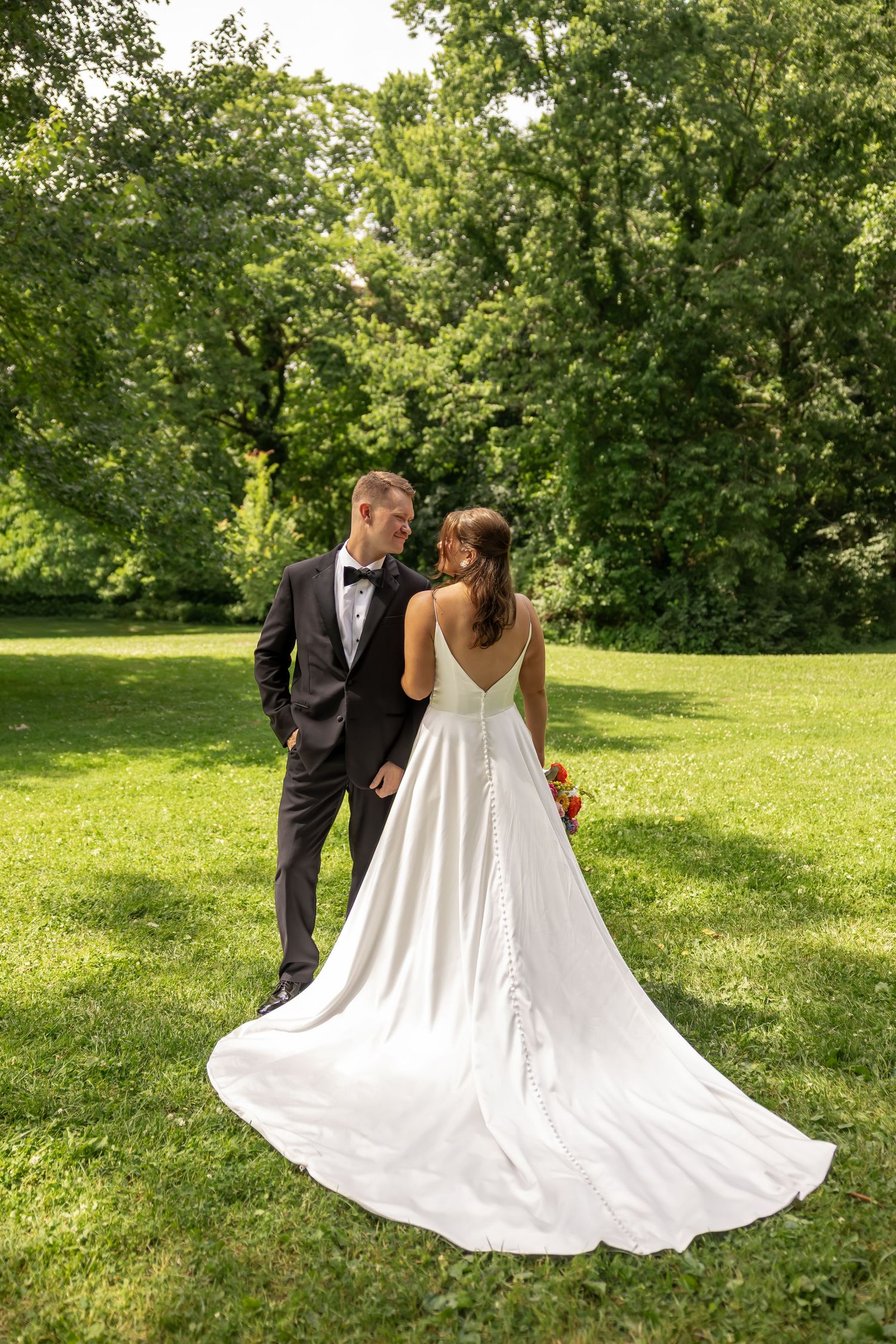 Bride and groom walking in lush green park, bride in elegant white gown with train, groom in black tuxedo.