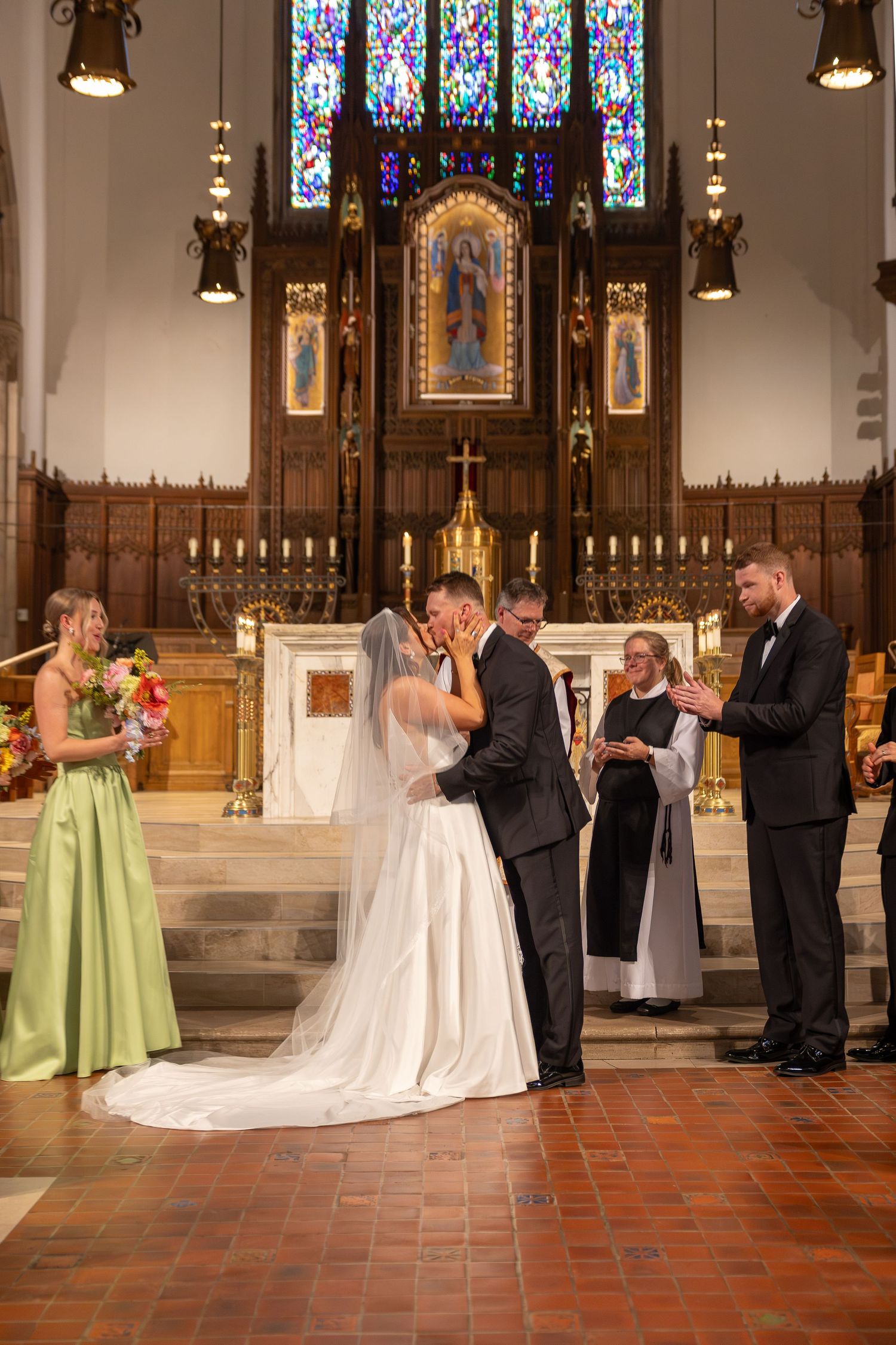 Bride and groom share first kiss at church altar with bridesmaid, groomsman, and officiant during wedding ceremony.