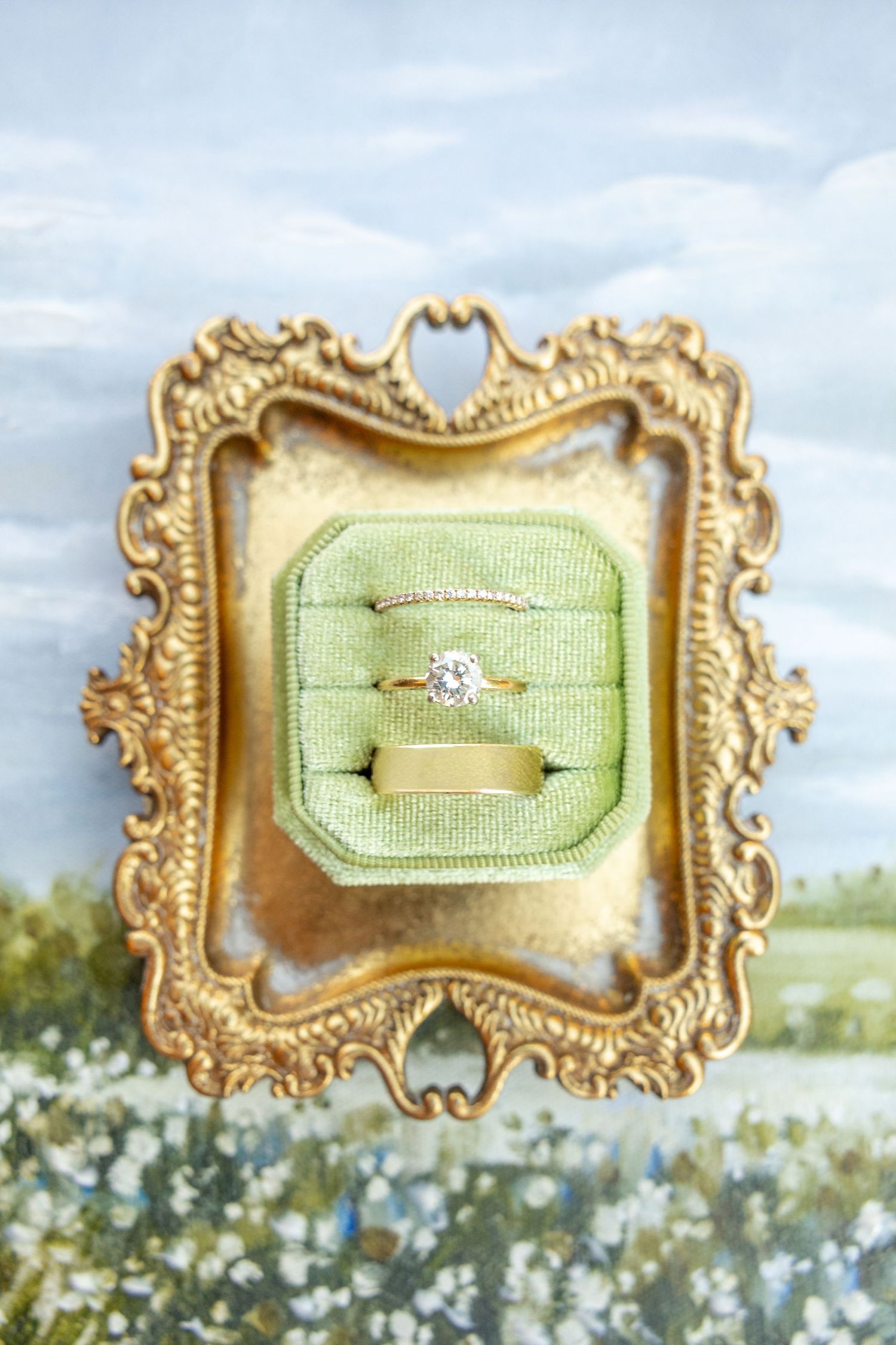 Gold engagement ring and wedding band displayed in a green velvet ring box within an ornate gold vintage frame.