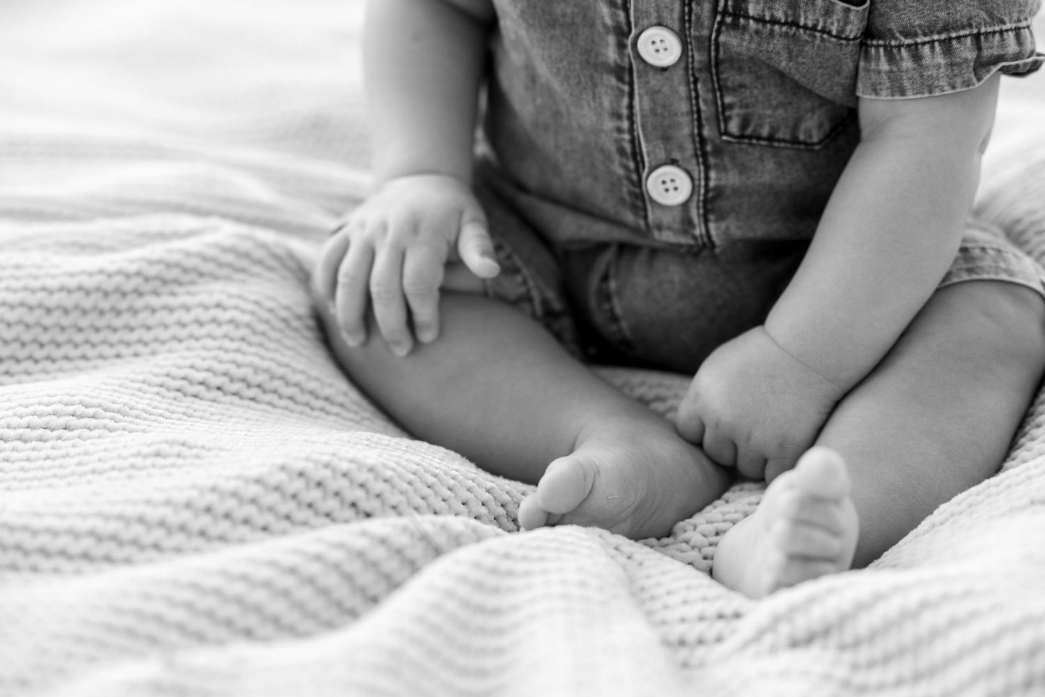 Black and white close-up of baby wearing denim overalls sitting on textured blanket.