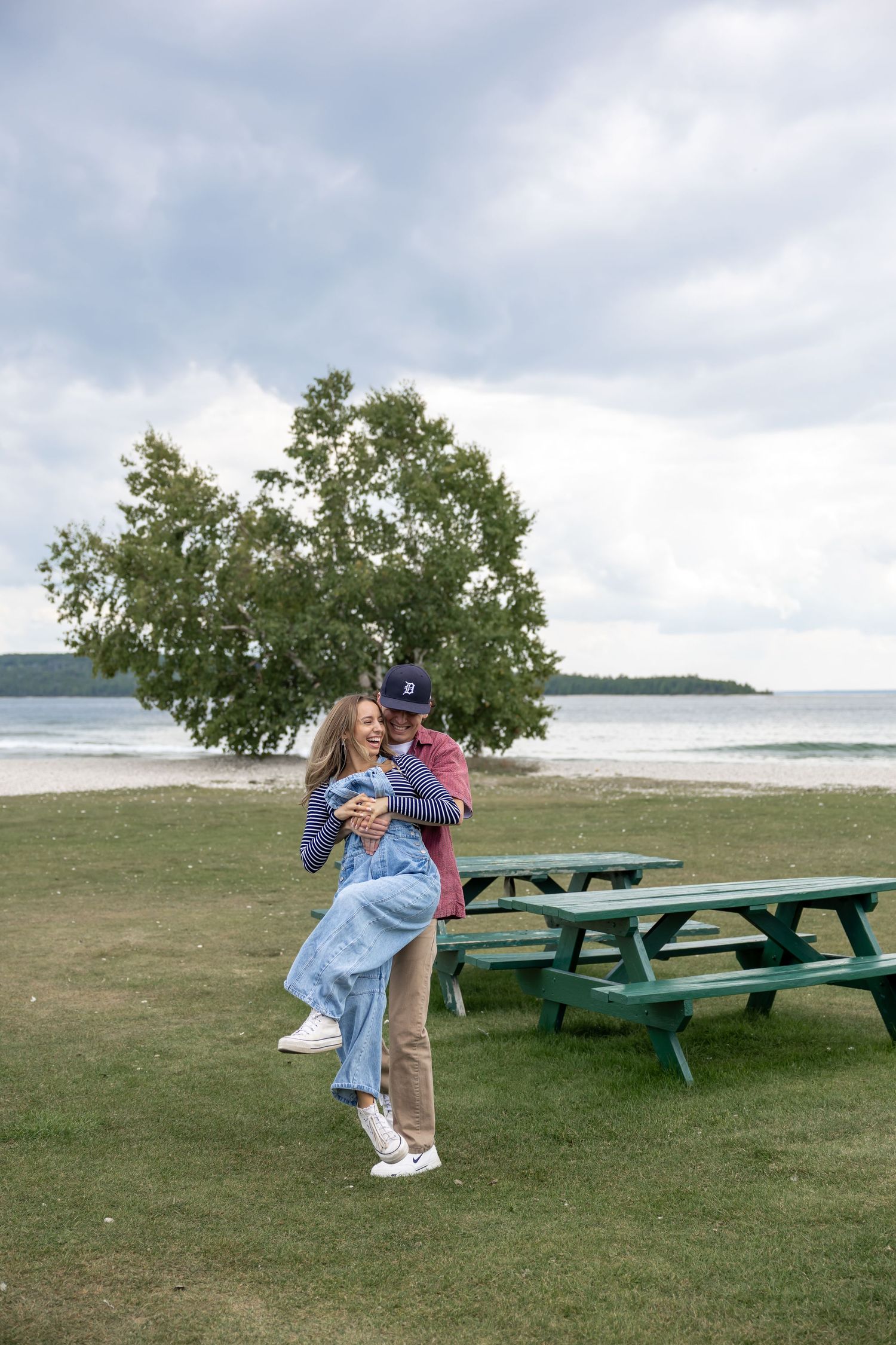 Two people embracing near picnic tables with the ocean and trees in the background.