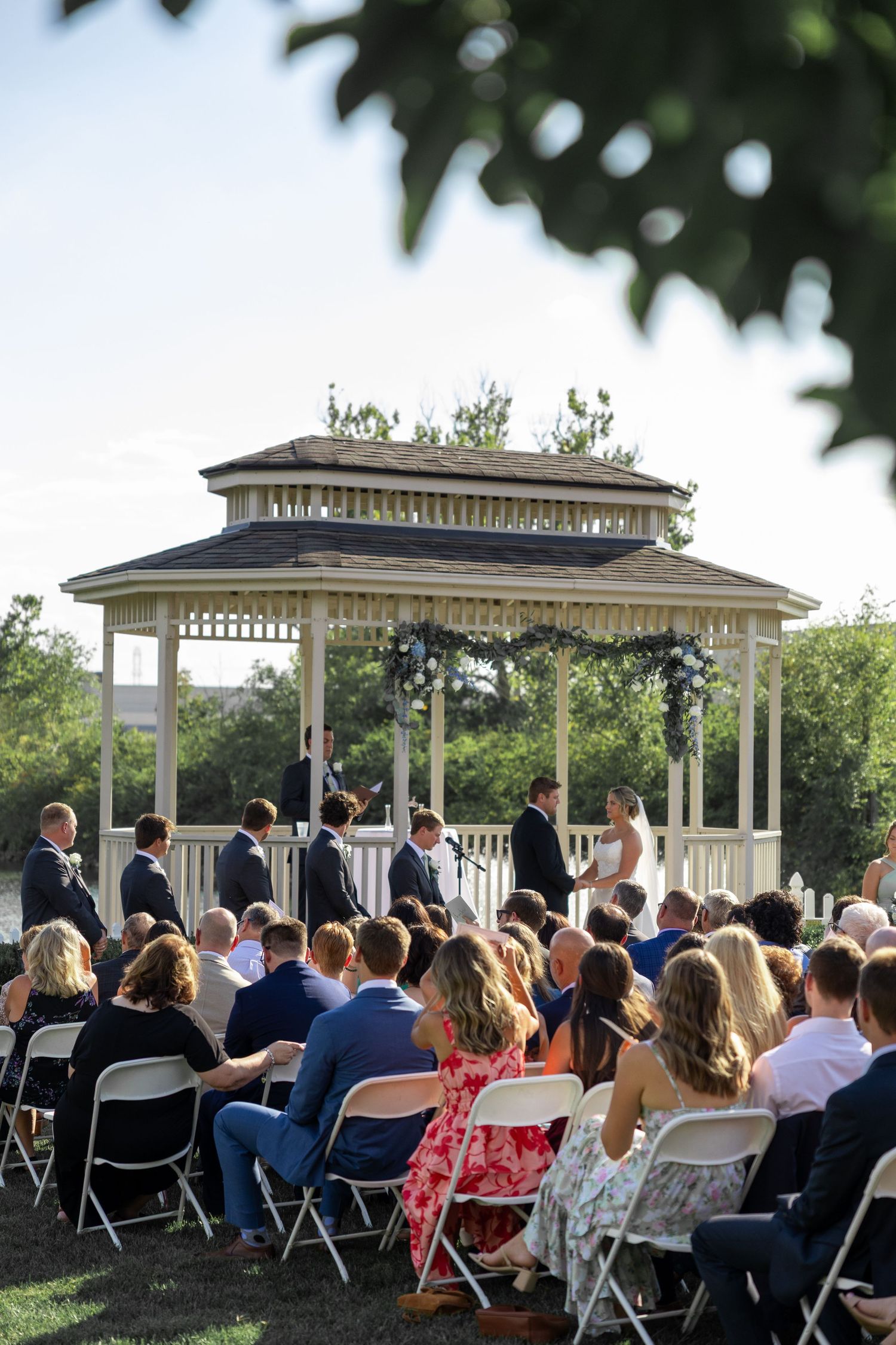 Outdoor wedding ceremony under a white gazebo with guests seated in rows of chairs.