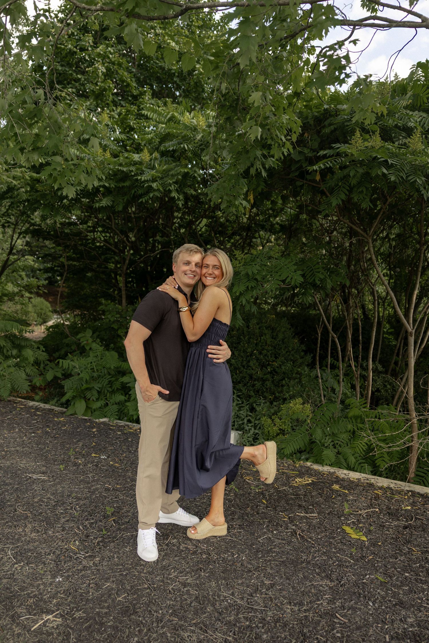 A smiling couple embraces outdoors, surrounded by lush green trees, woman in blue dress, man in black shirt.
