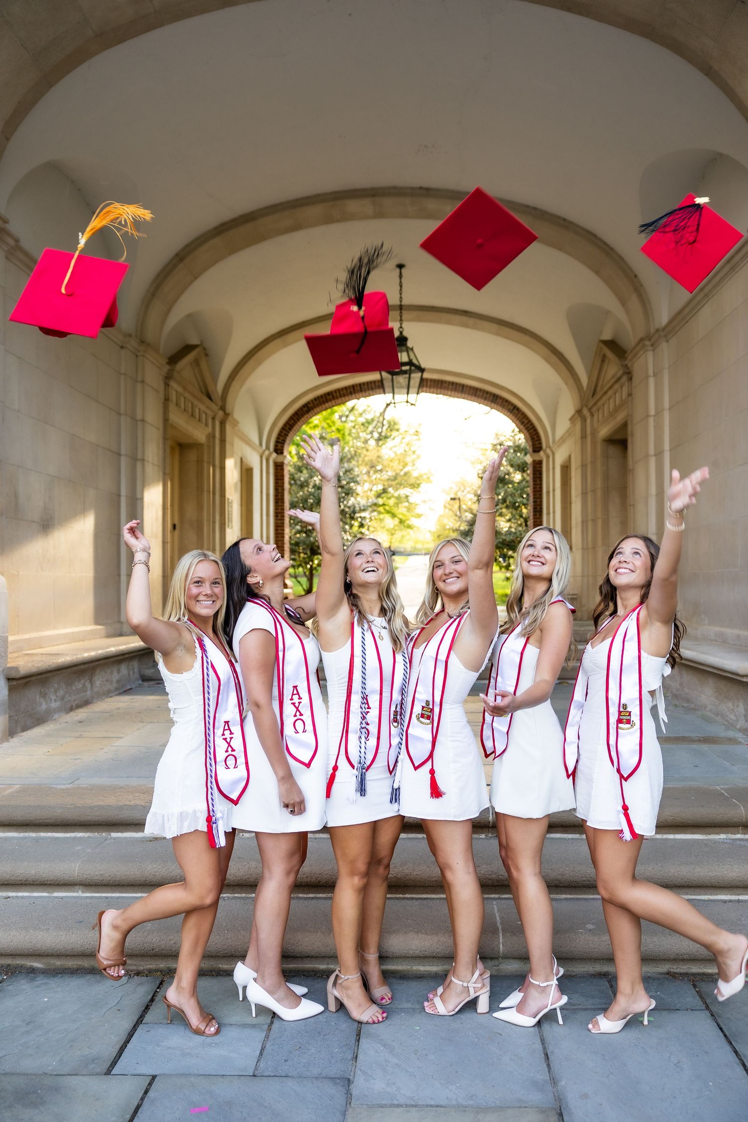 Graduation photo of students in white dresses and red sashes posing under an archway.