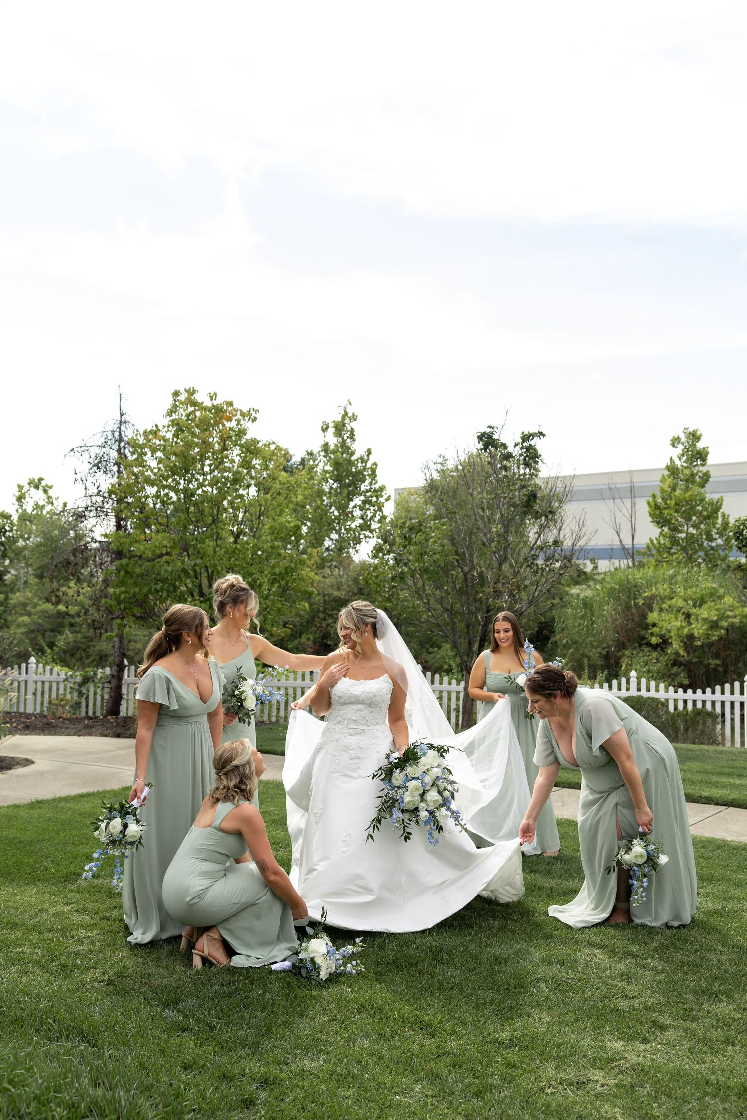 Bridesmaids in sage green dresses help arrange wedding dress on grass.