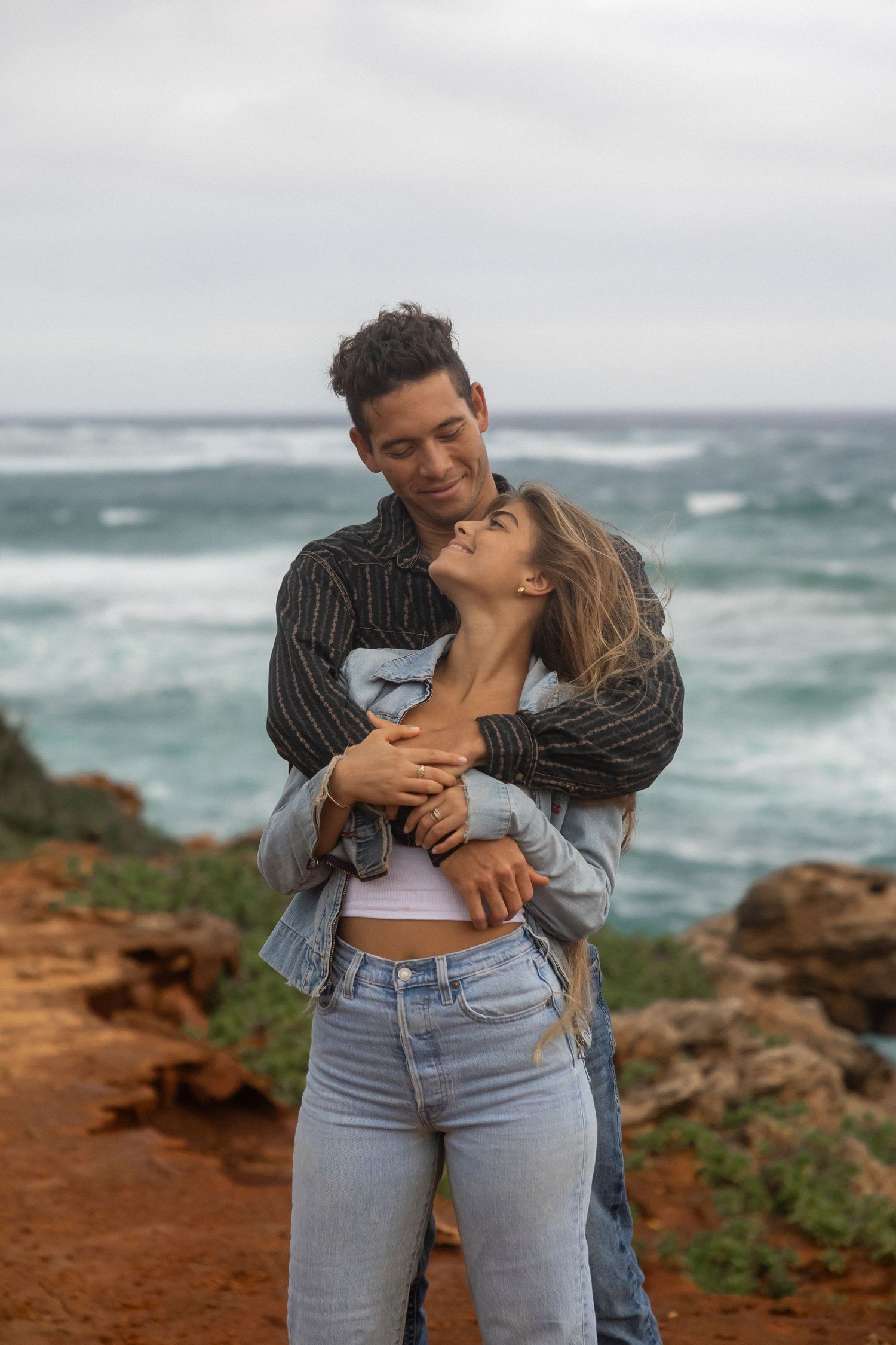 Romantic couple embraces on a dramatic coastal cliff with crashing waves in the background.