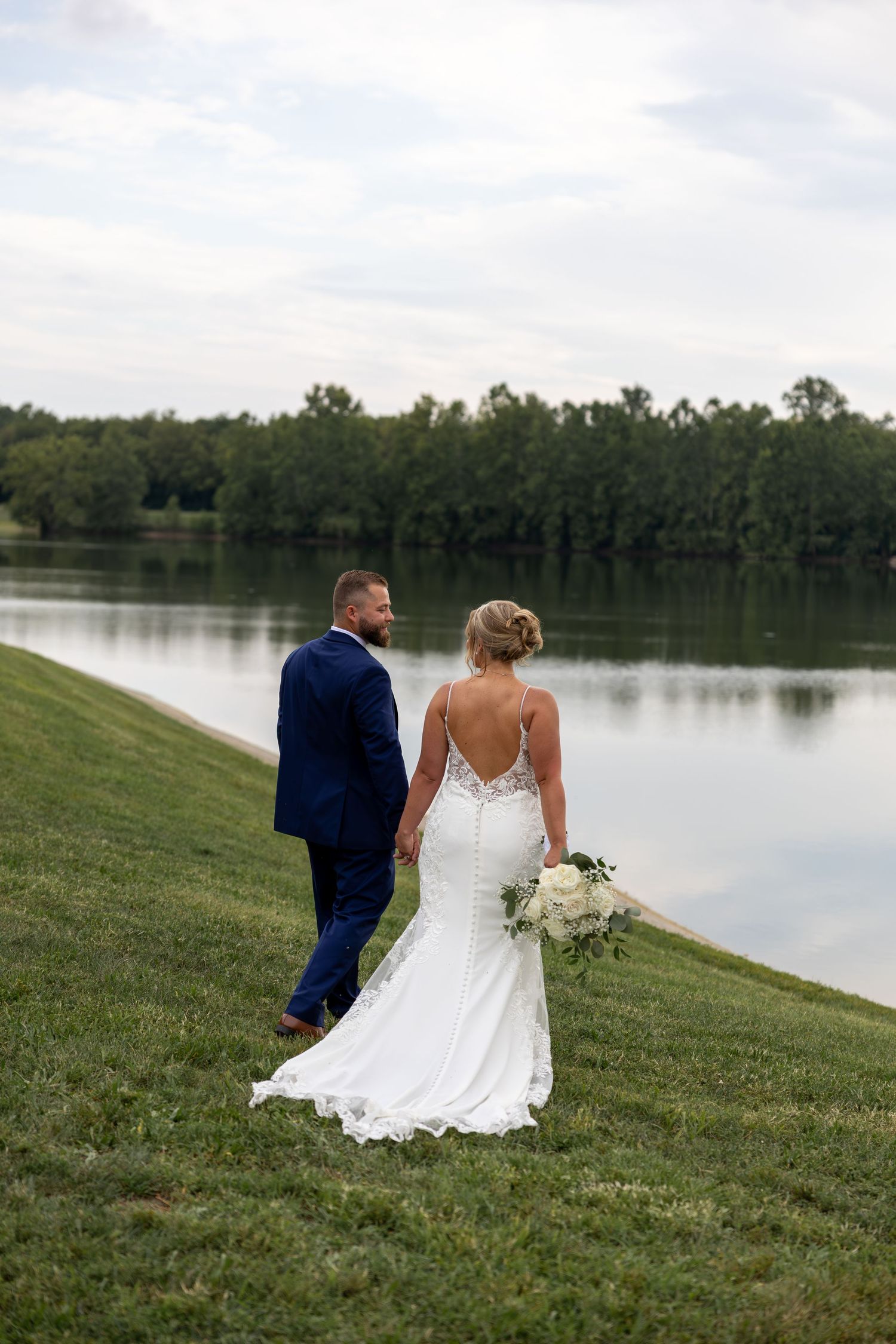 Couple in wedding attire sitting on grassy hill overlooking serene lake.