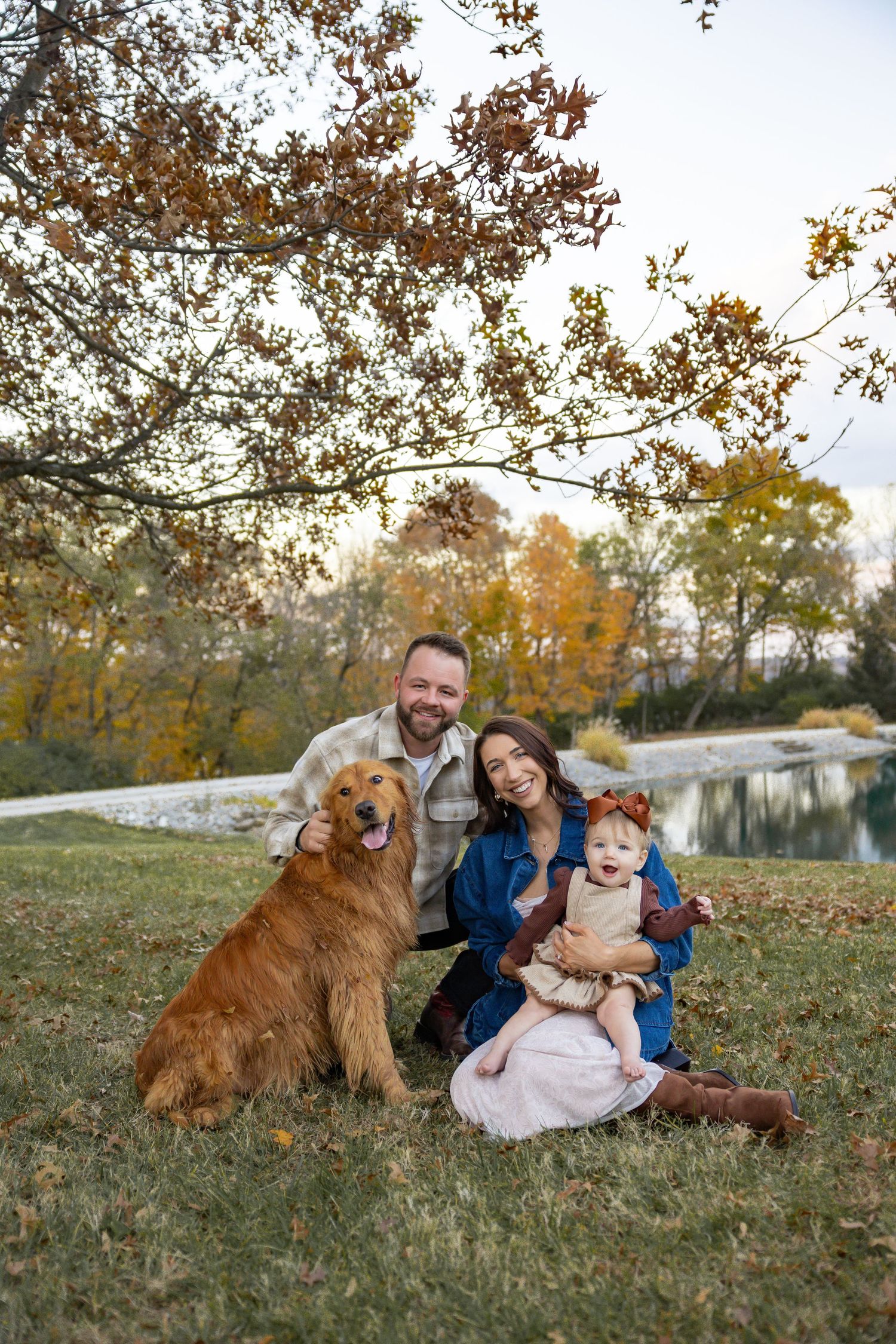 Family portrait with two golden retrievers relaxing on autumn grass near a pond.