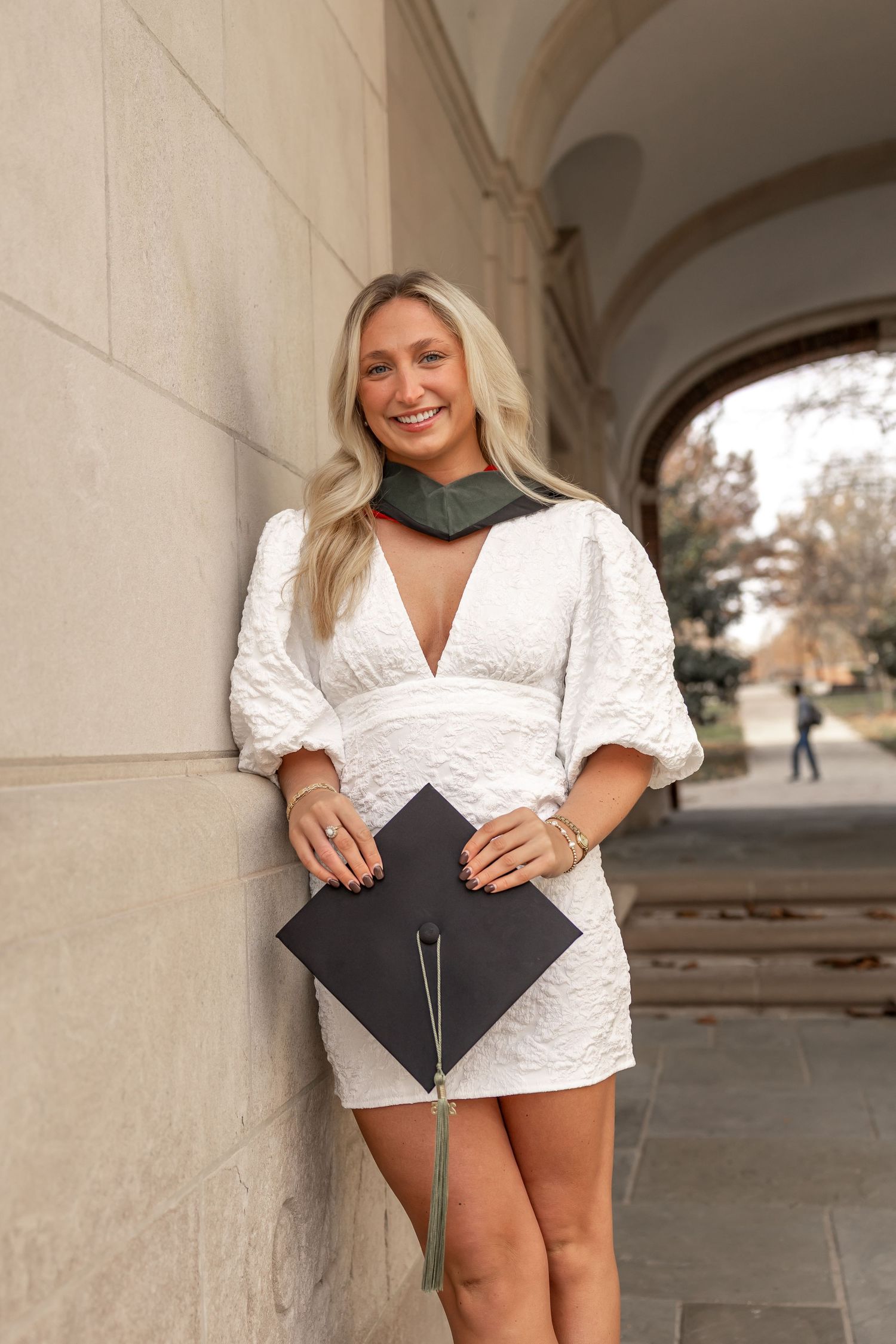 Graduate in white dress posing with diploma against stone archway background.