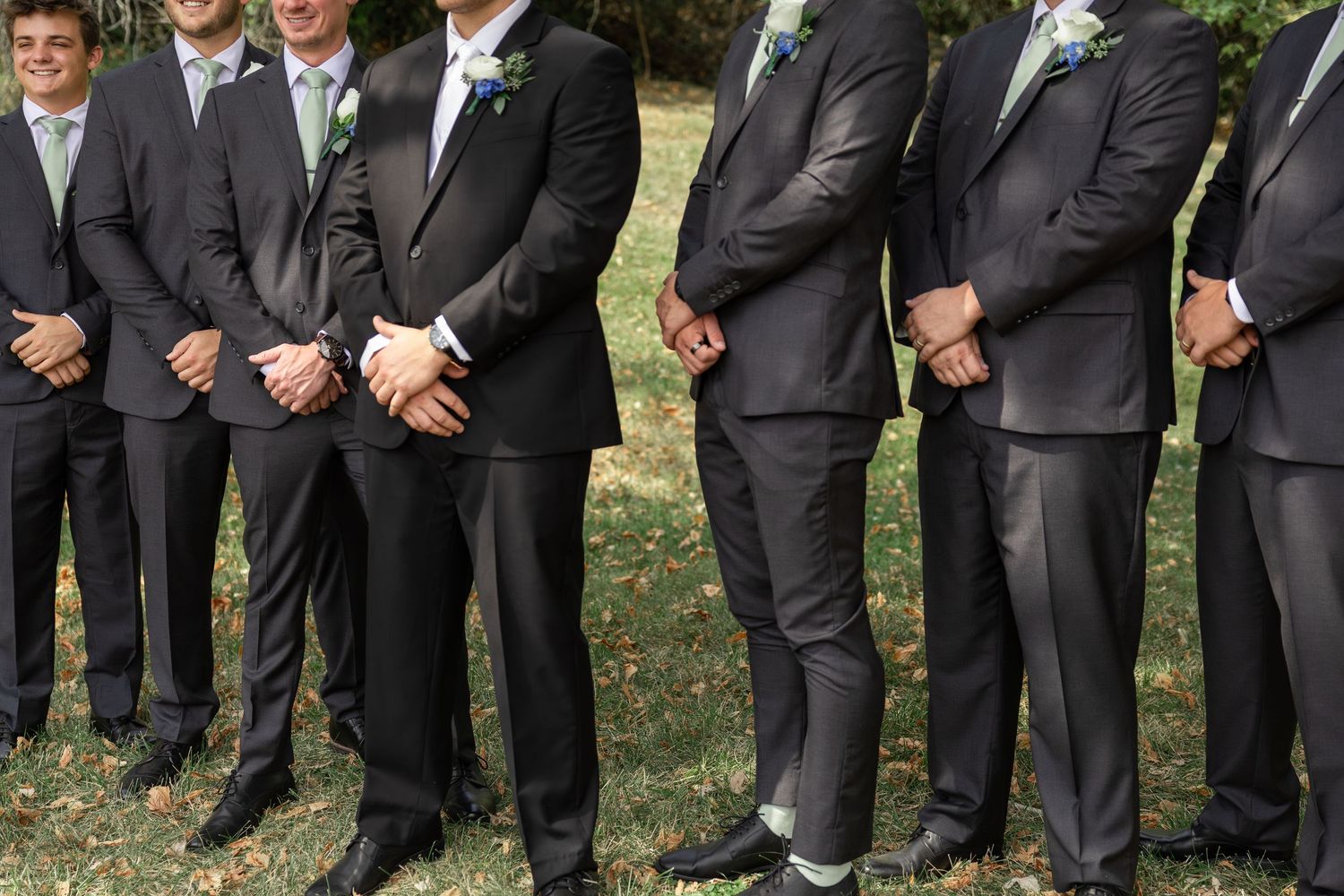 Row of groomsmen in black suits and ties standing together at outdoor wedding.