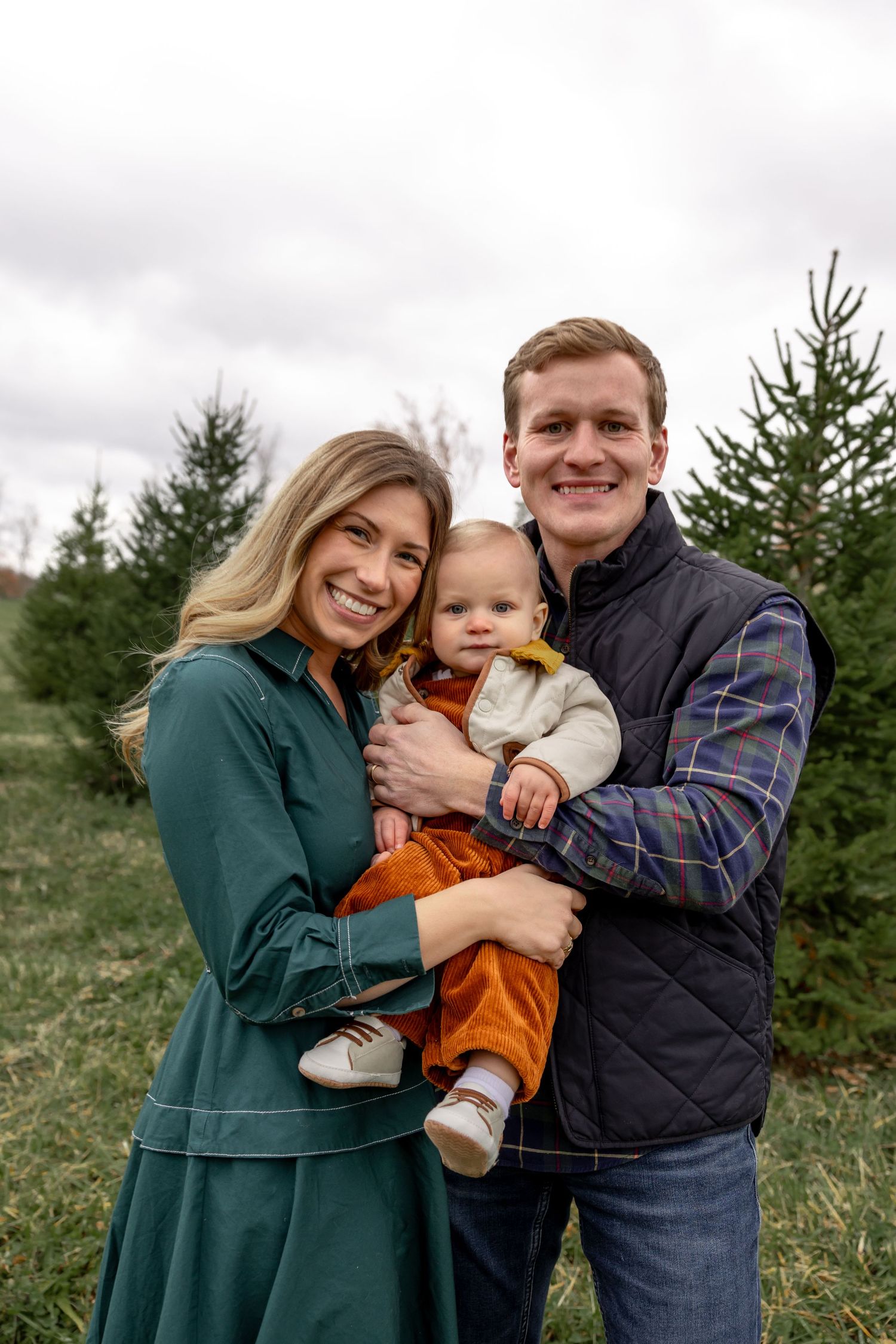 Family of three poses together among evergreen trees on a cloudy day.