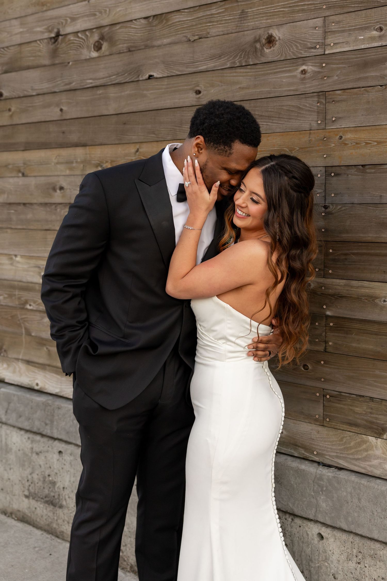 A bride in a white gown and groom in a black tuxedo share a romantic moment against a rustic wood wall.