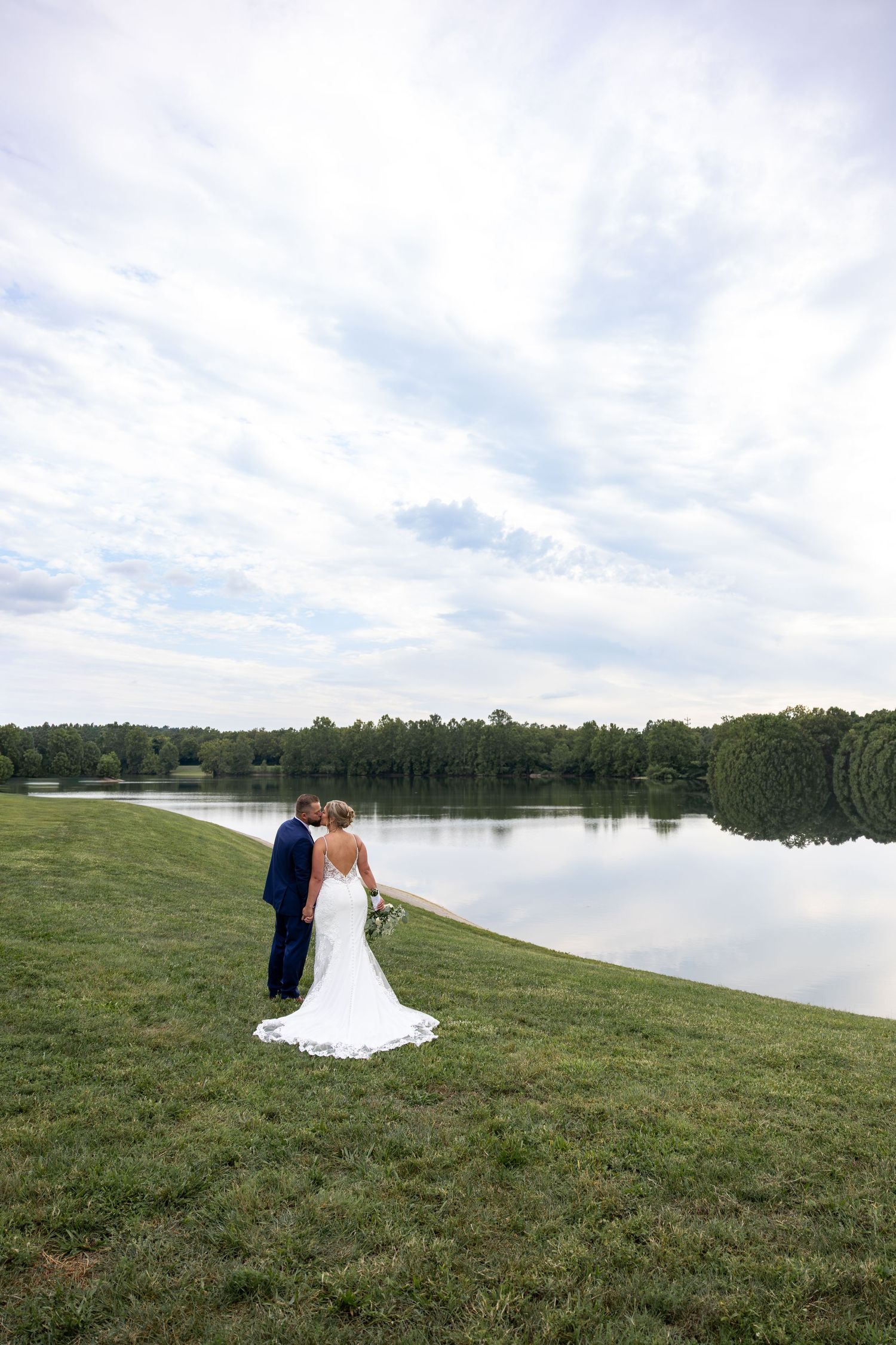 A couple in wedding attire poses by a serene lakeside on a cloudy day.