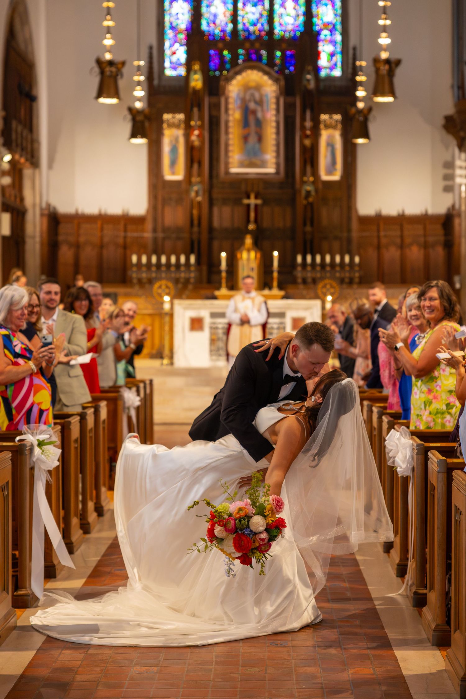 Bride and groom share a romantic dip kiss in church aisle after wedding ceremony with guests applauding.