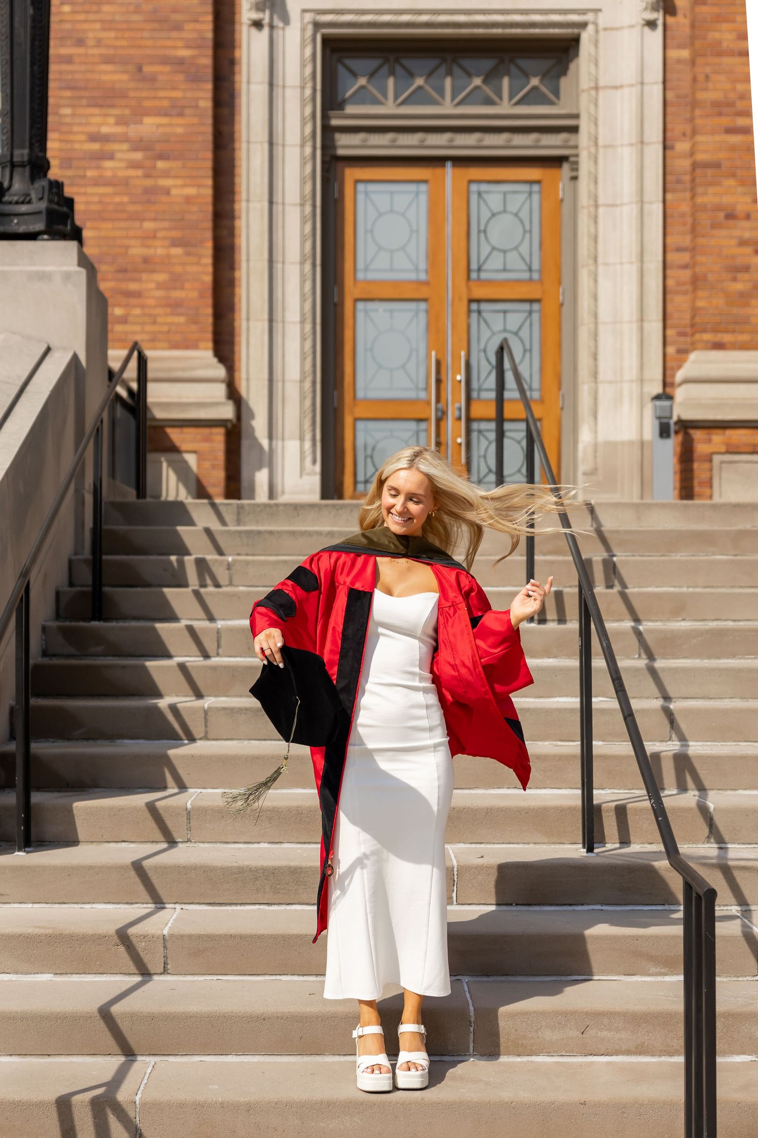 Smiling blonde graduate in red robe and white dress poses on stone steps outside a university building.